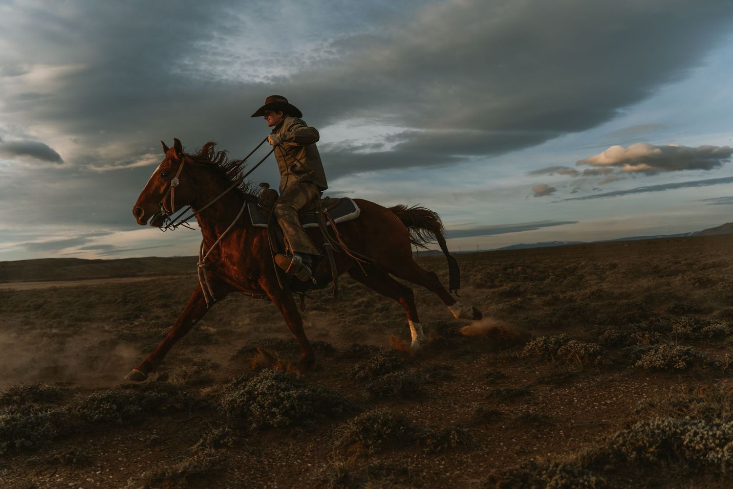A cowboy rides a galloping brown horse across a vast open plain under a dramatic cloudy sky at dusk.