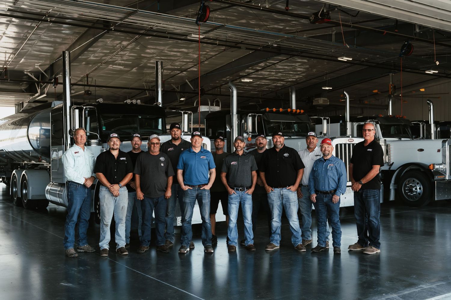 Group of truck drivers posing in front of semi-trucks inside a large commercial trucking facility.