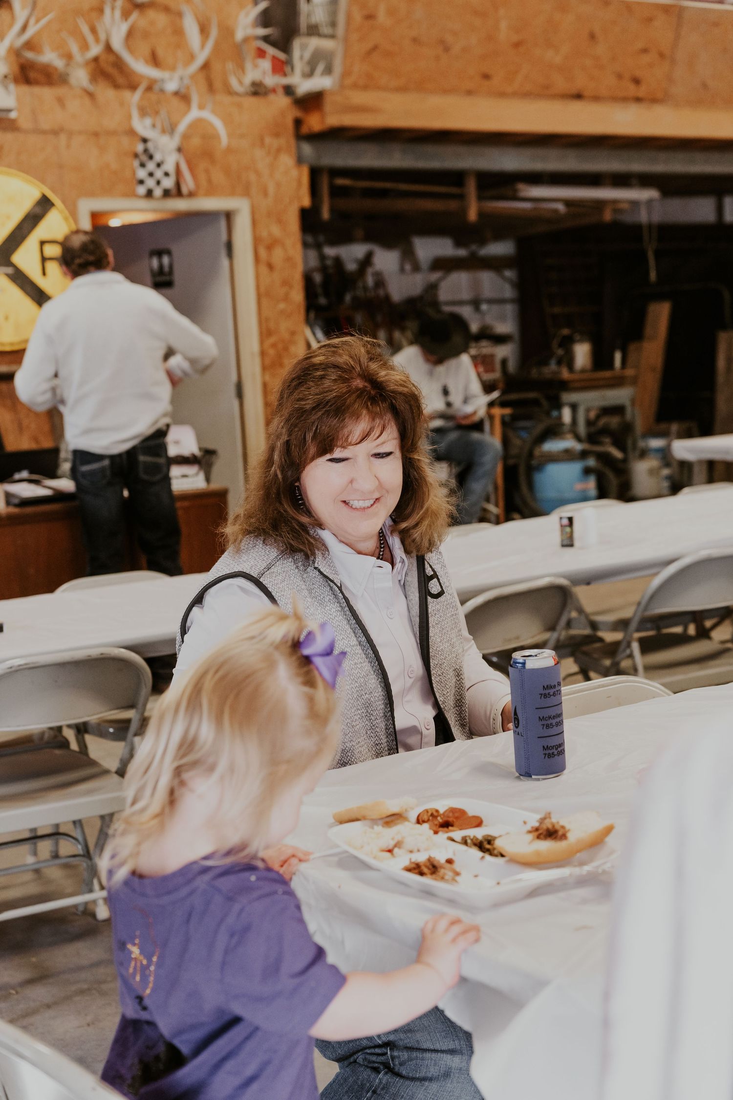 Woman smiling at young girl over a food tray at an indoor community event with folding tables and chairs.