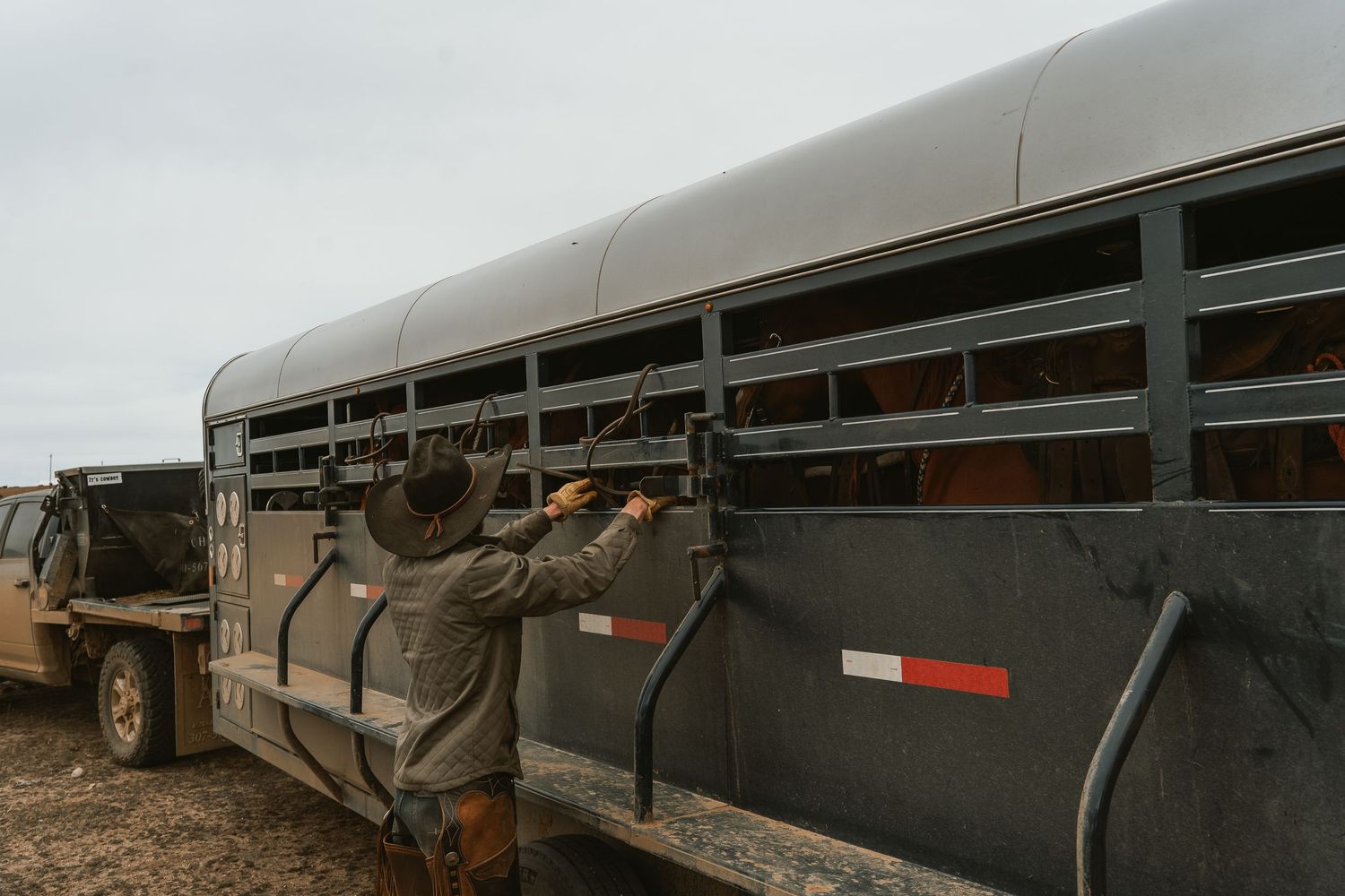 A cowboy in a hat secures latches on a dark livestock trailer on a cloudy day.
