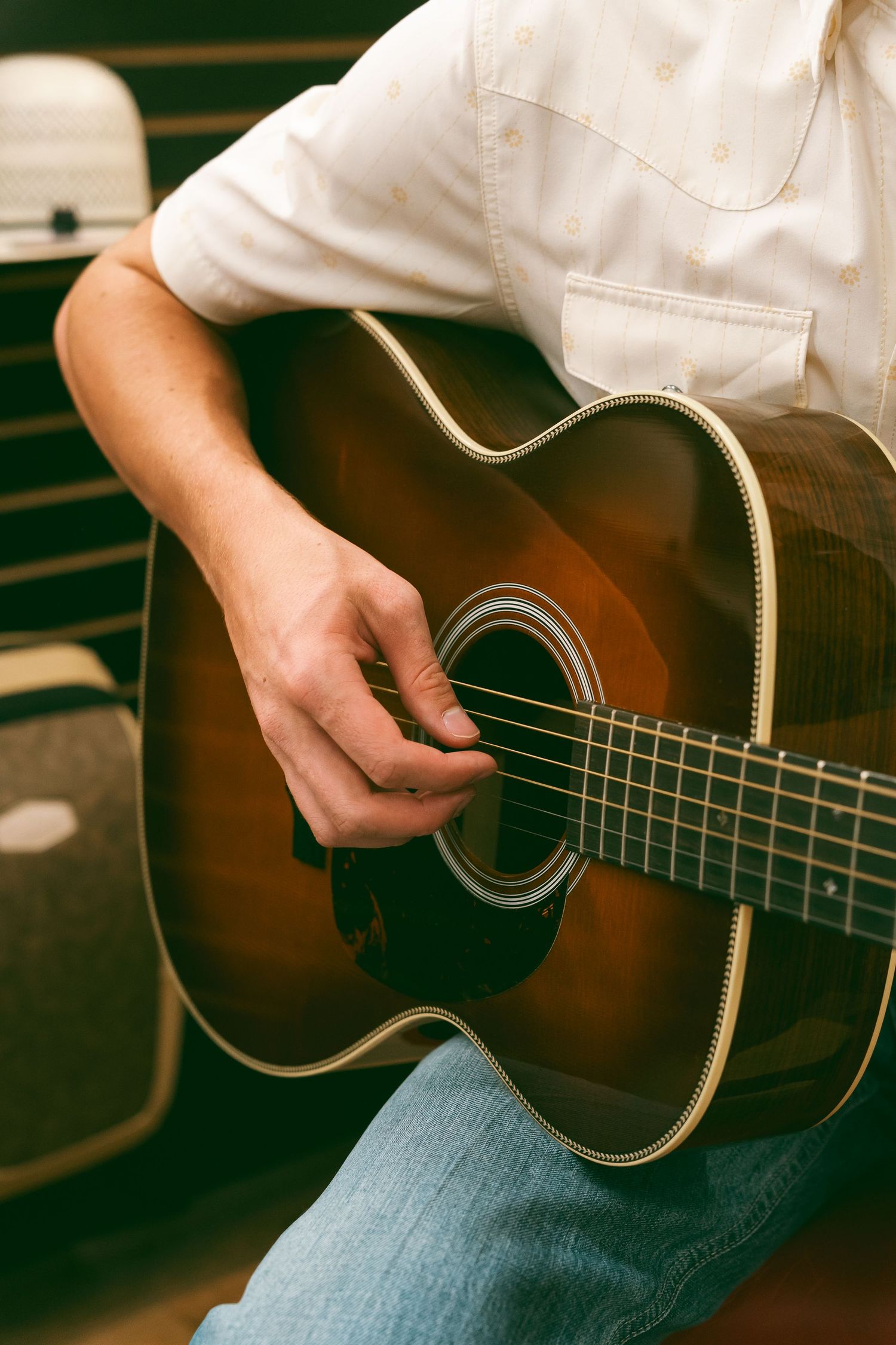 Close-up of a man in a white shirt strumming an acoustic guitar while seated, wearing blue jeans.