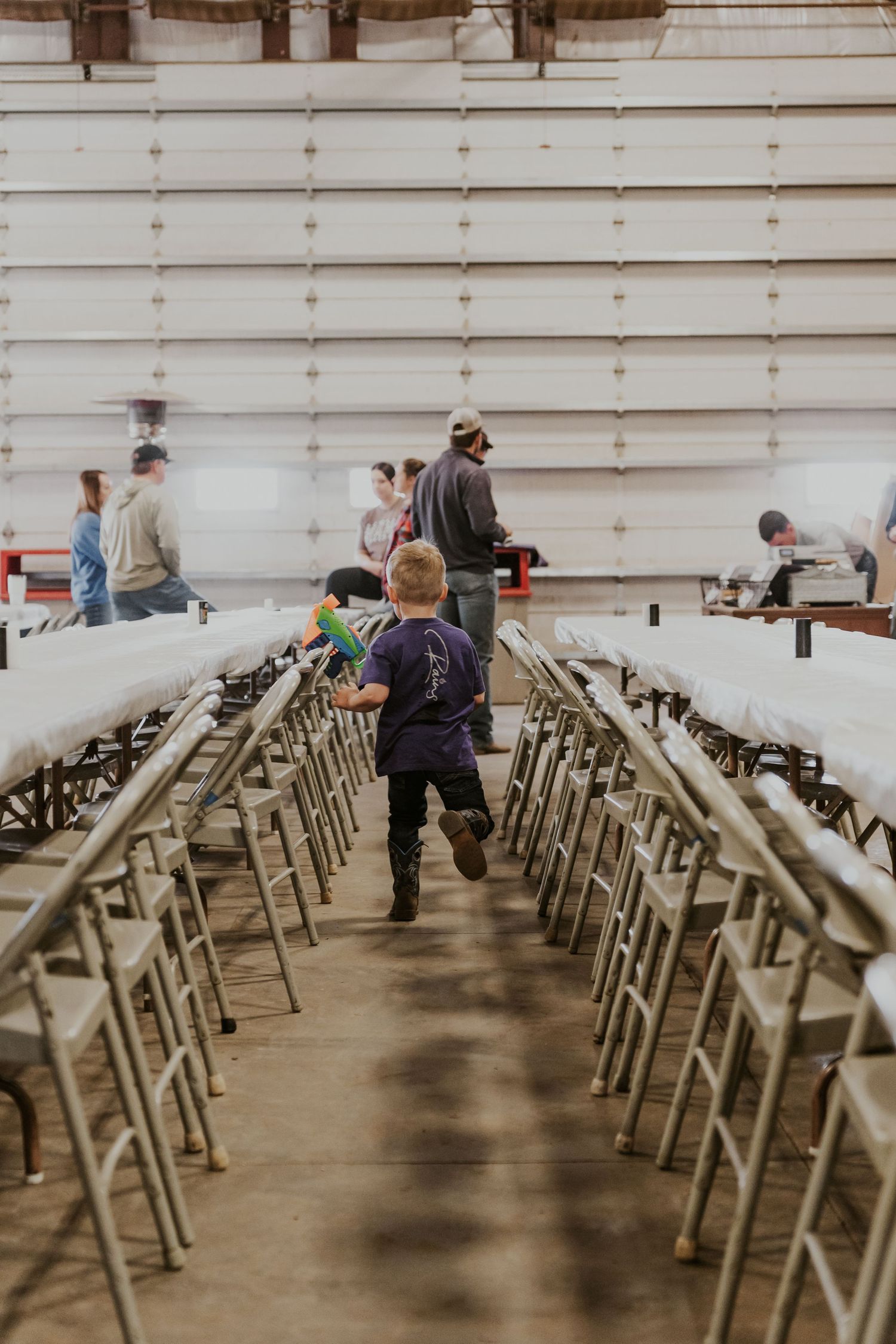 Young boy running between rows of folding tables and chairs inside a large event hall.