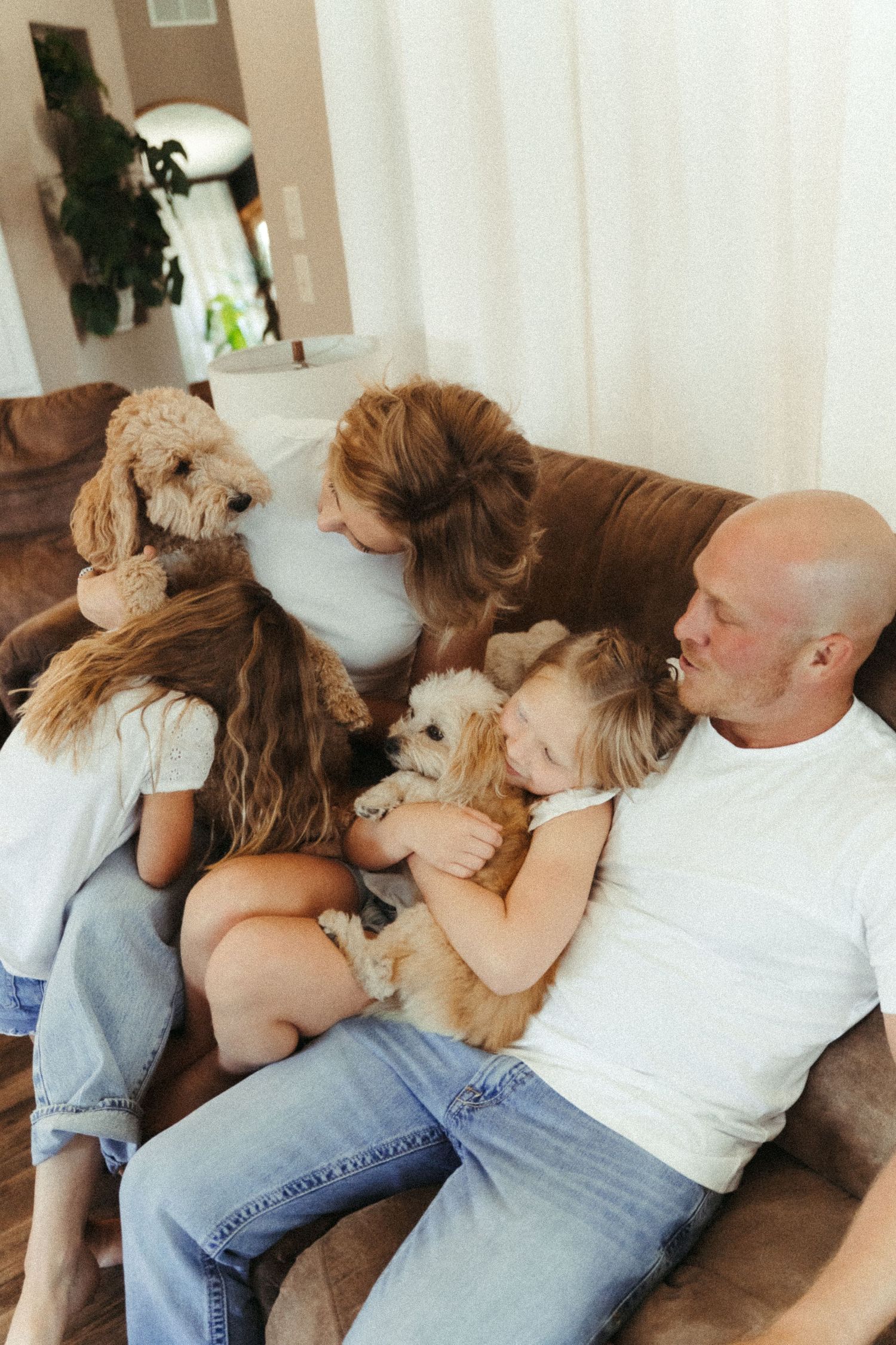 Family with two girls and fluffy dogs cuddling on brown sofa at home.