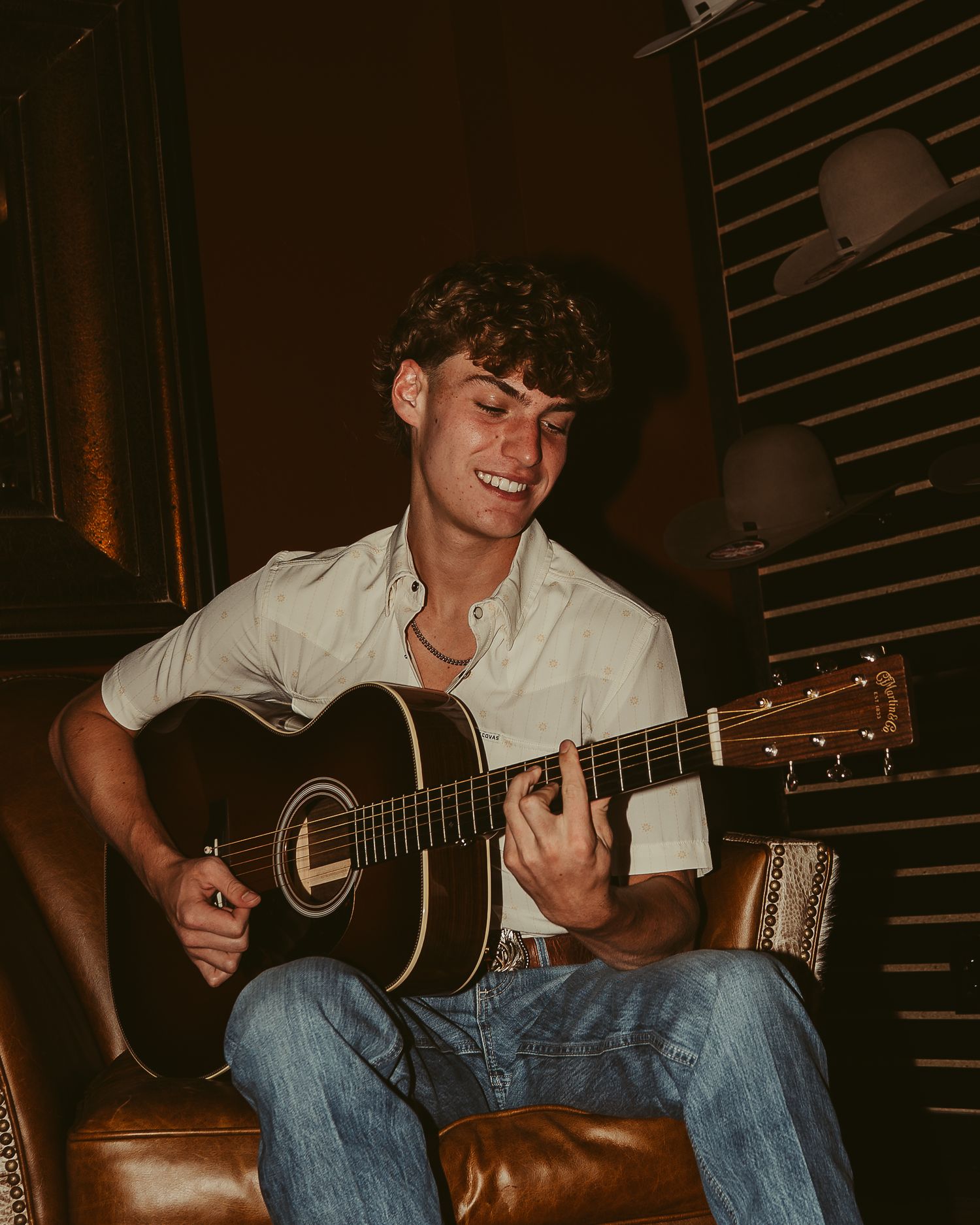 Young man smiling while playing acoustic guitar, seated on leather chair in warm ambient lighting.