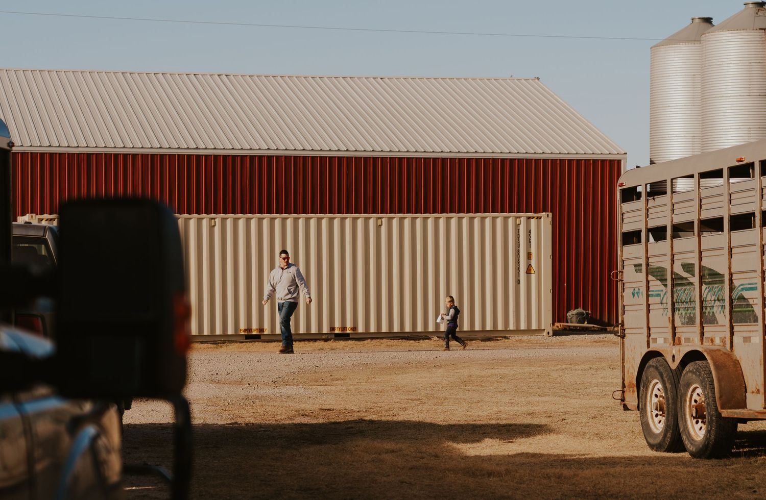 Red barn with shipping containers and livestock trailer on a rural farm with two people visible outside.