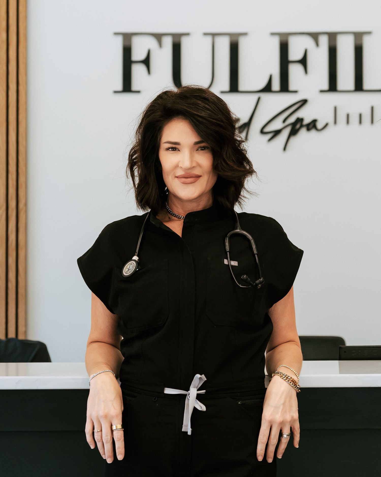 A woman in black scrubs with a stethoscope stands smiling at a med spa reception desk.