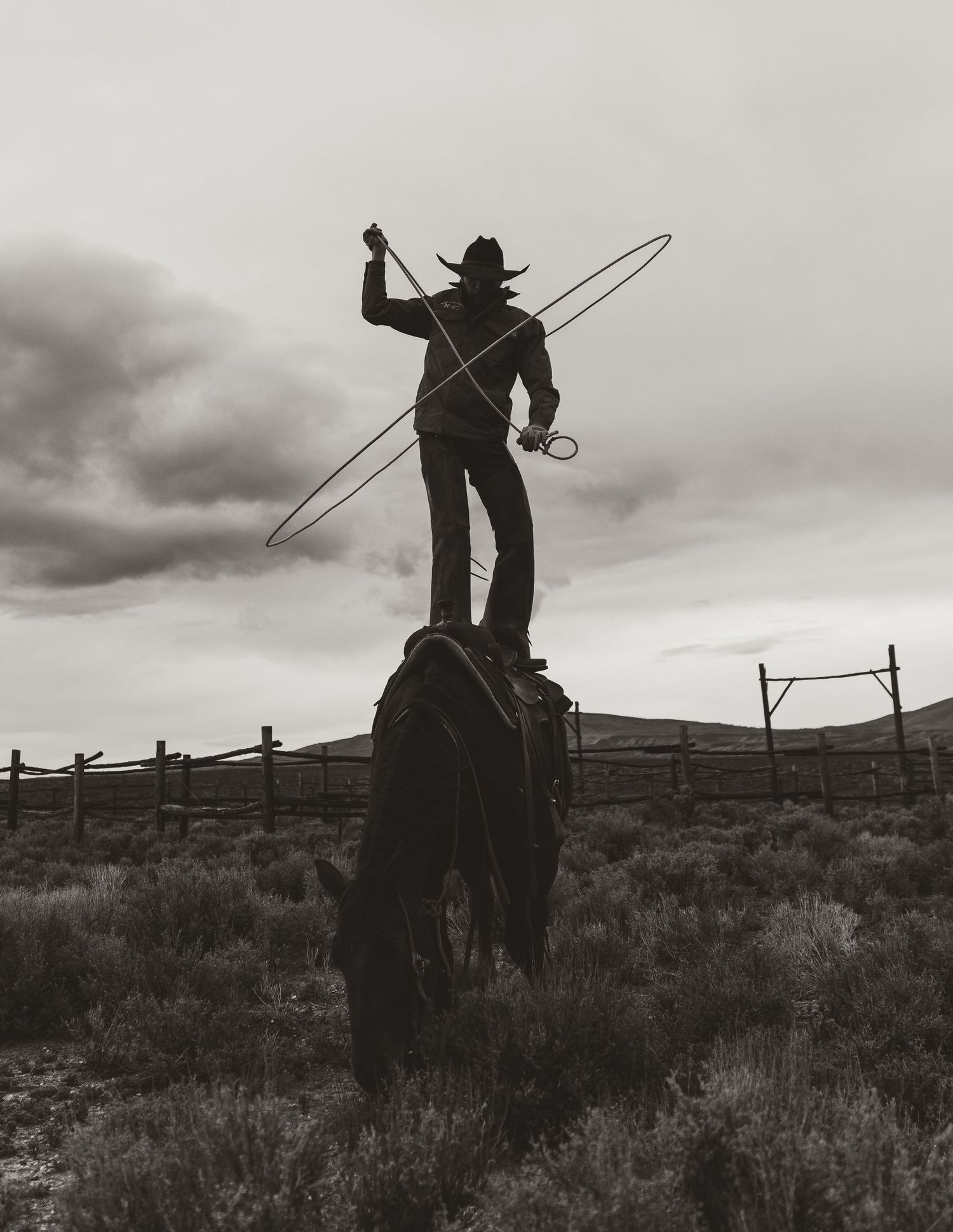 Black and white photo of a cowboy standing on horseback, swinging a lasso in an open field under a cloudy sky.