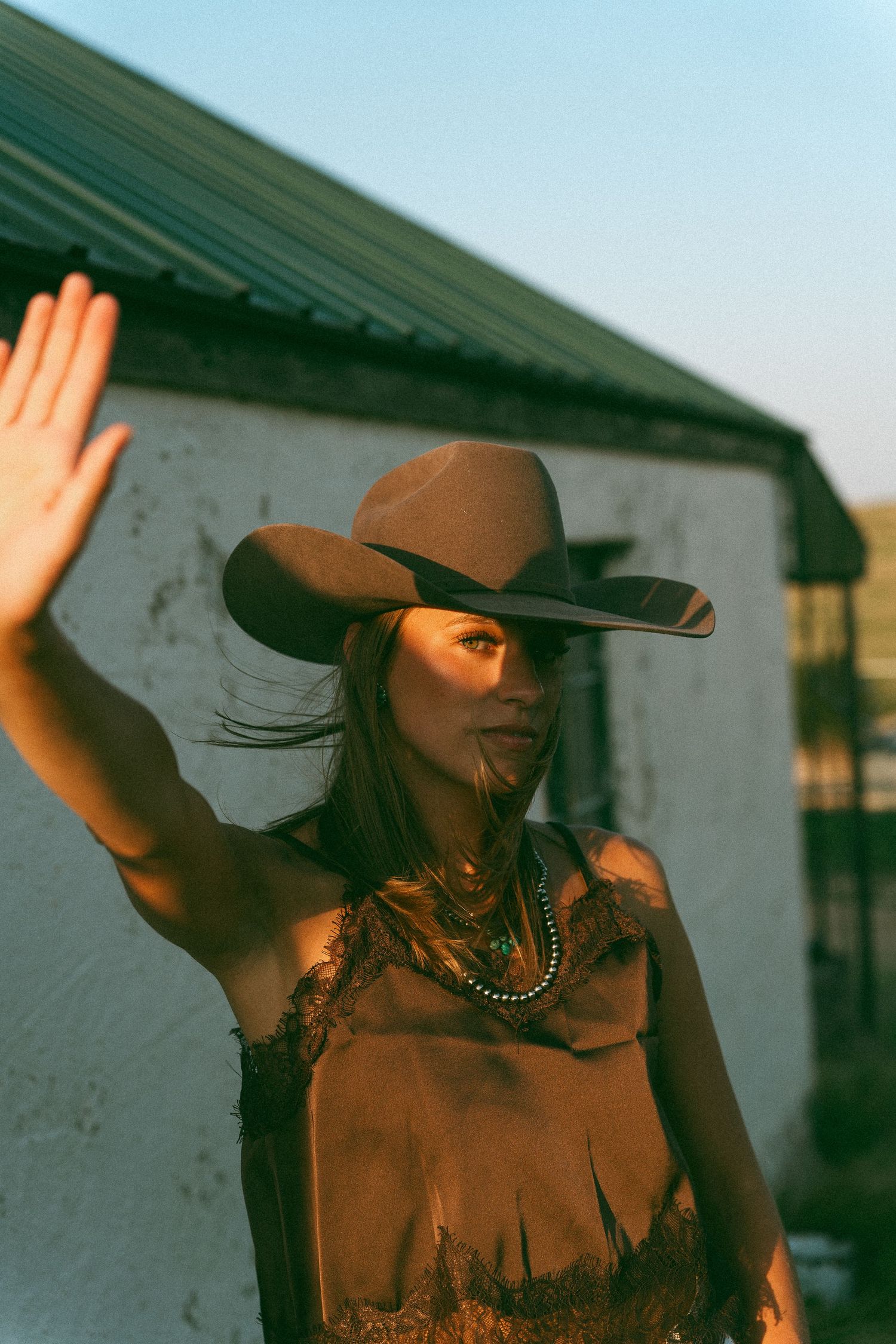 Woman in brown cowboy hat and fringe top raising hand in golden sunlight near white building.