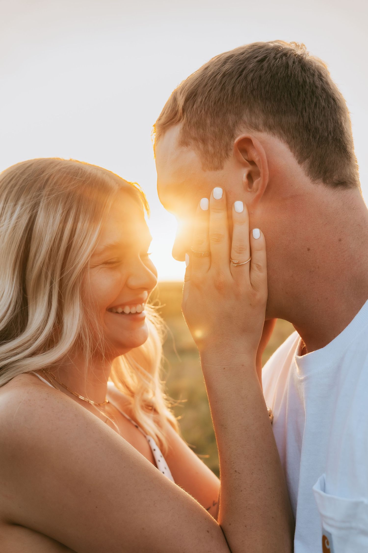 Couple sharing a romantic moment at golden hour sunset, woman smiling with engagement ring visible on her hand.