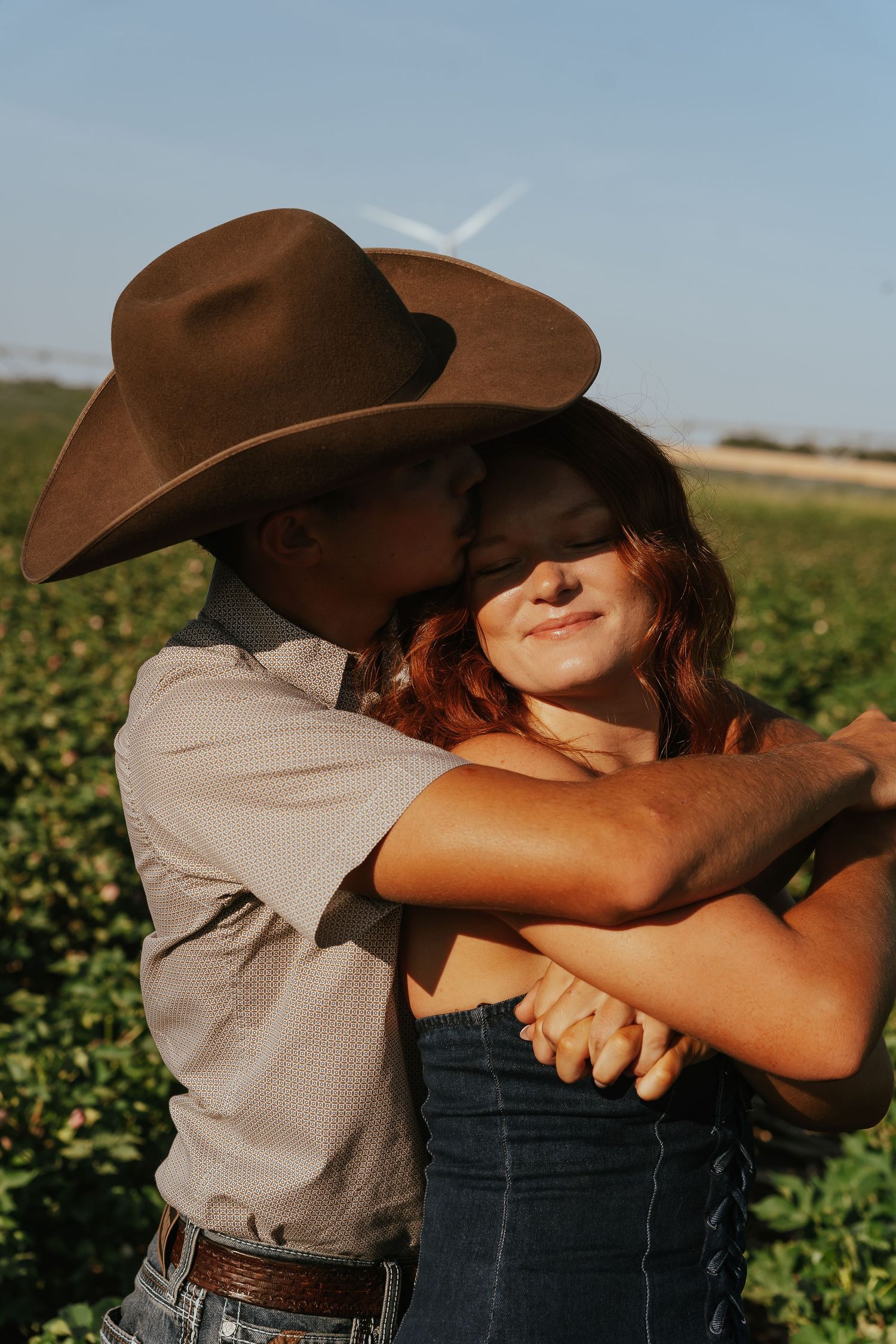 Couple embracing in a green field, man in cowboy hat kissing woman's cheek during golden hour.