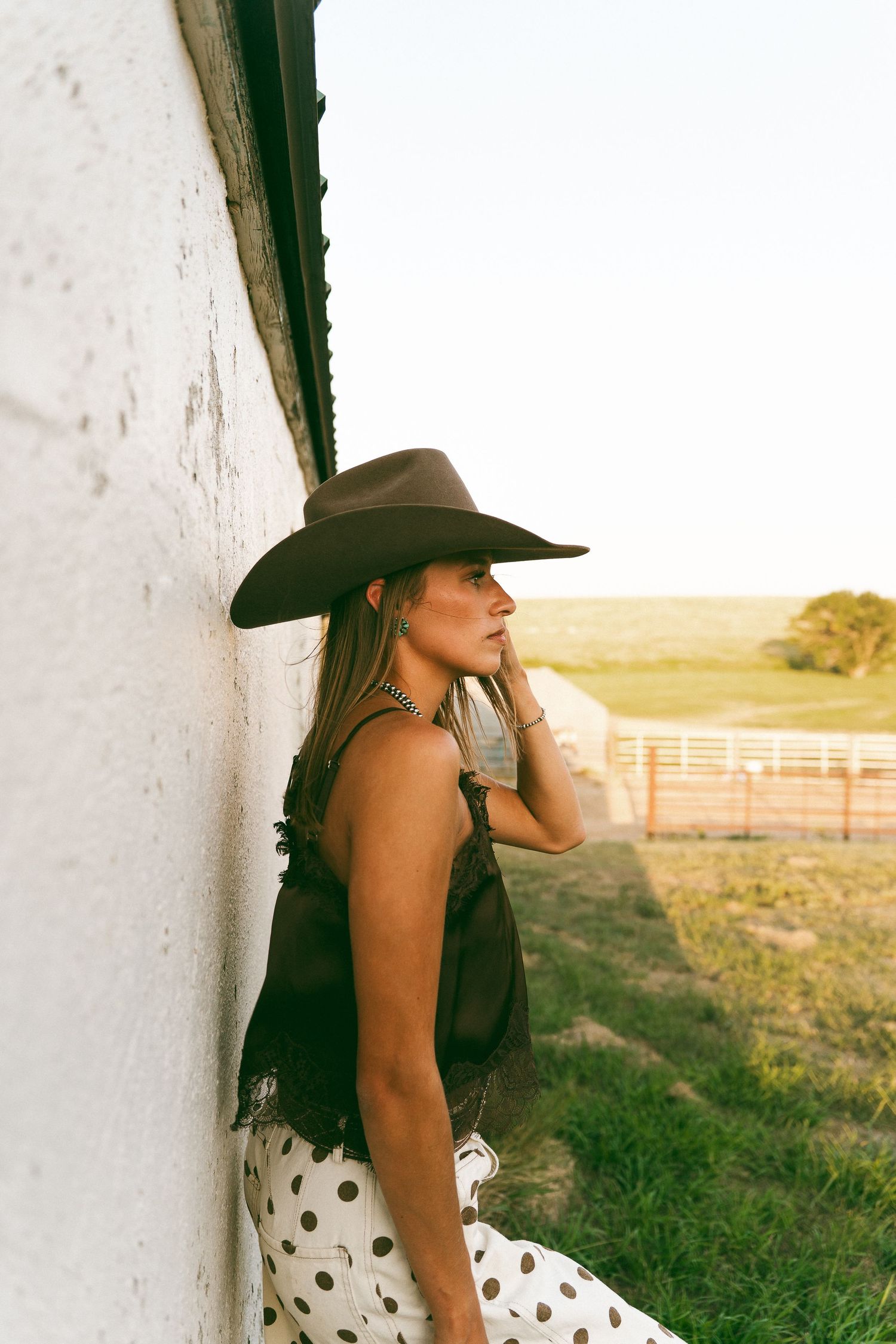 Woman in wide-brim cowboy hat and polka dot skirt leaning against white barn wall at golden hour.