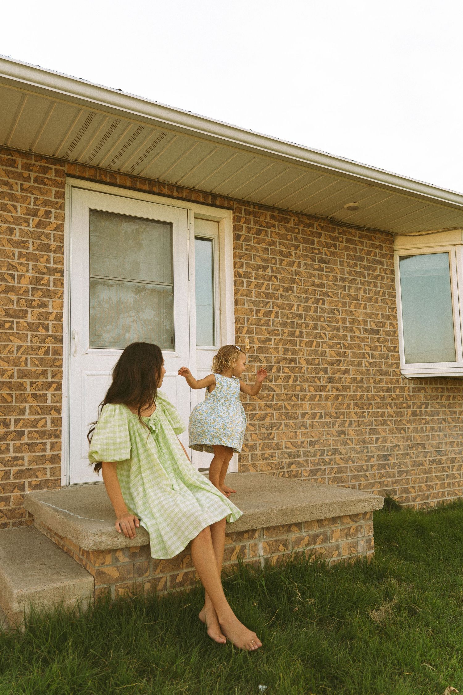 Two young girls in summer dresses playing on brick house front steps outdoors.
