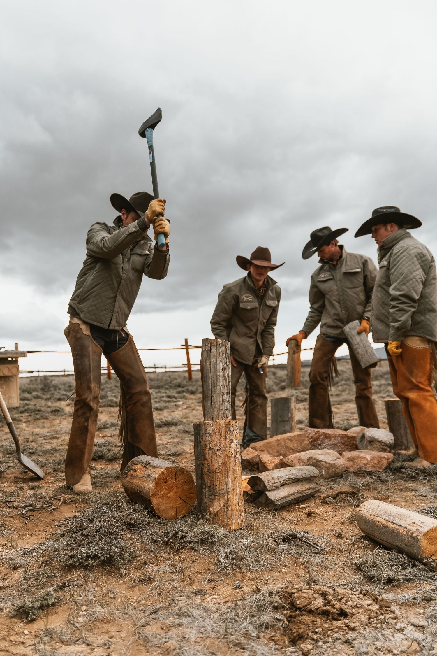 Cowboys in hats chop firewood with an axe on a cloudy day in a rustic outdoor setting.