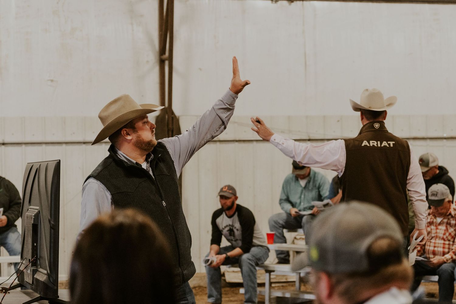 Two cowboys in Ariat vests high-five at a western ranch event inside a large barn.