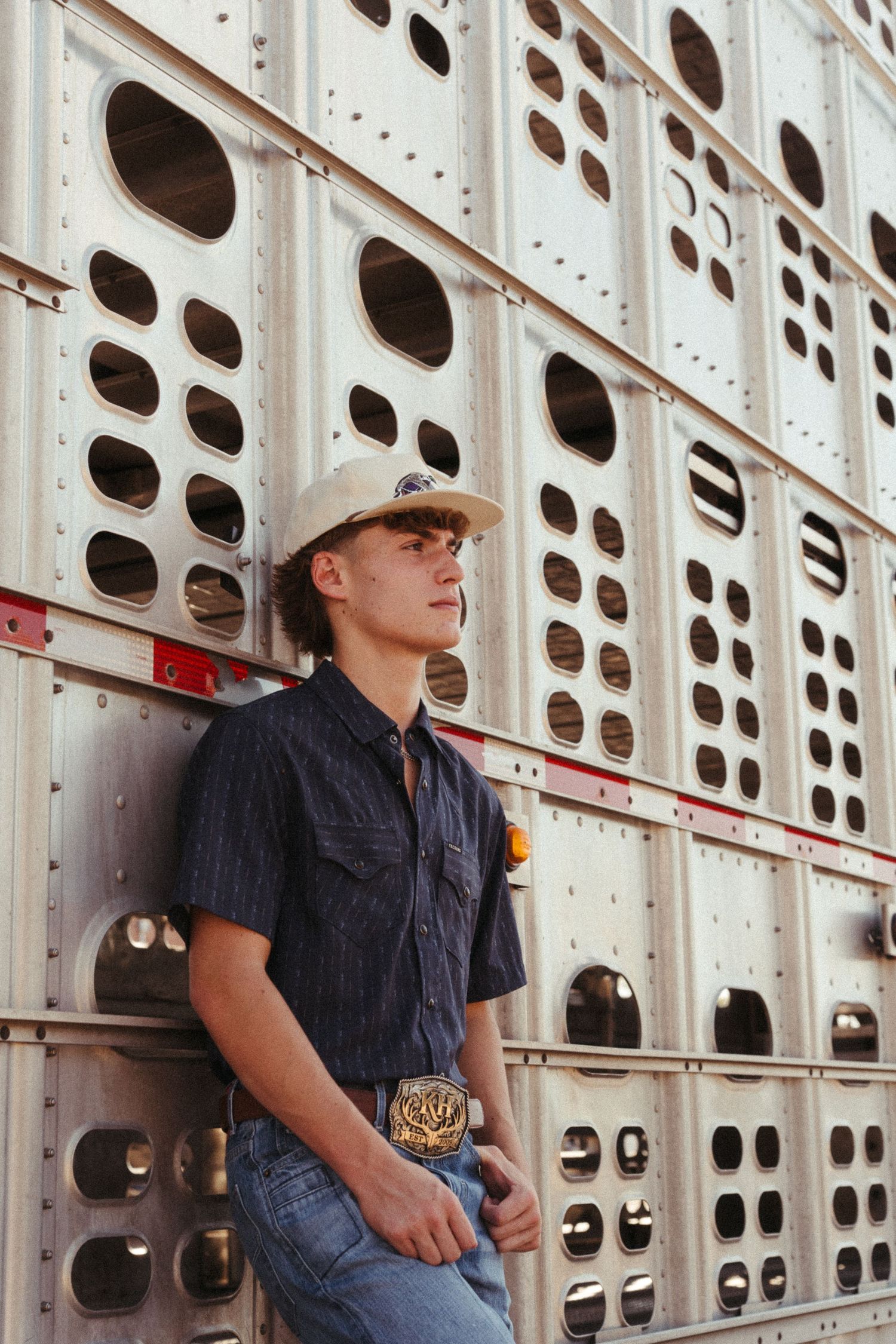 Young cowboy in cap and belt buckle leans against silver livestock trailer in jeans and dark shirt.