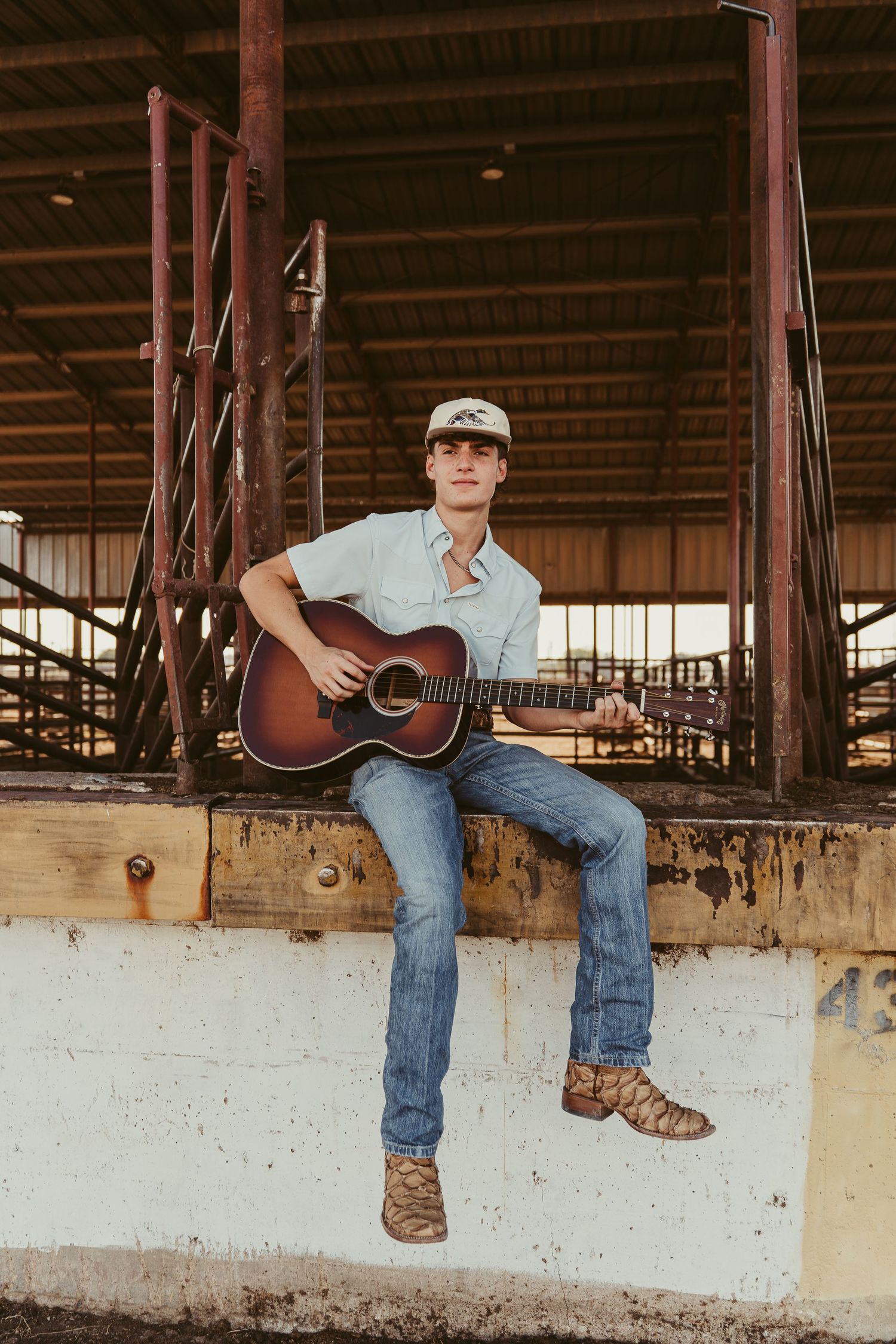 Young cowboy plays acoustic guitar while sitting on arena fence wearing boots, jeans, and cap.
