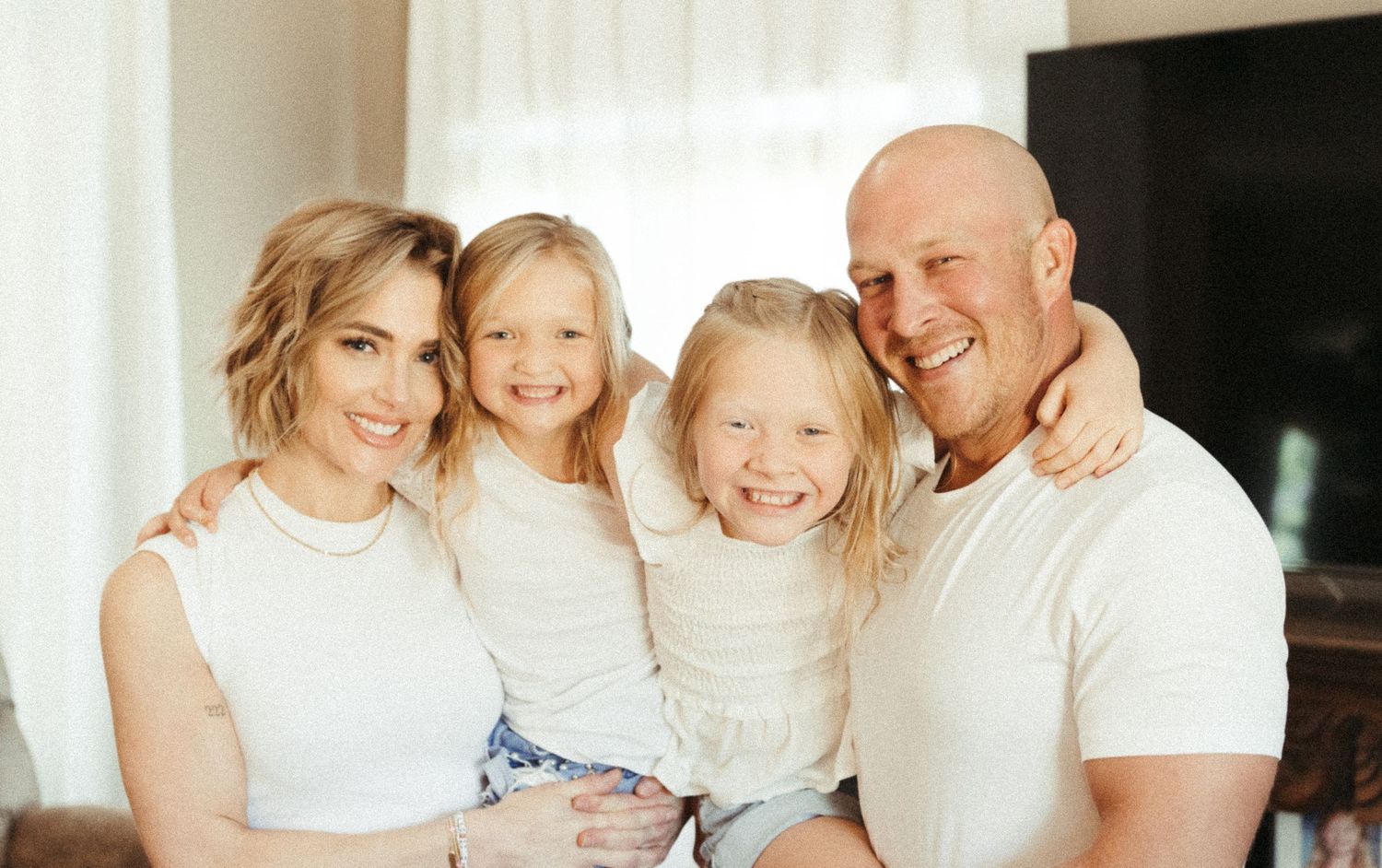 Happy family of four smiling together, featuring two young girls and their parents wearing white tops indoors.