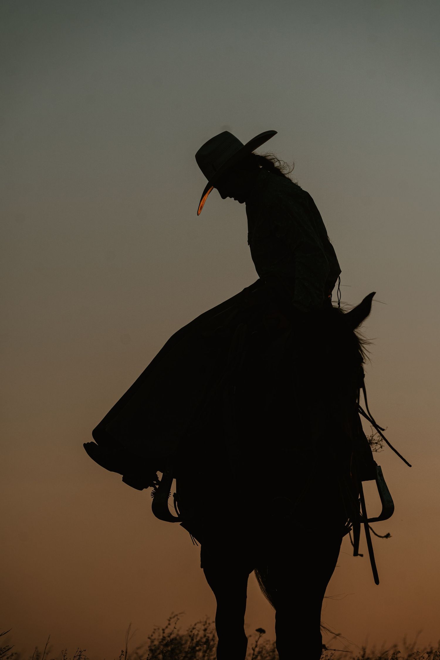 Silhouette of cowboy on horseback against warm orange and gray sunset sky.