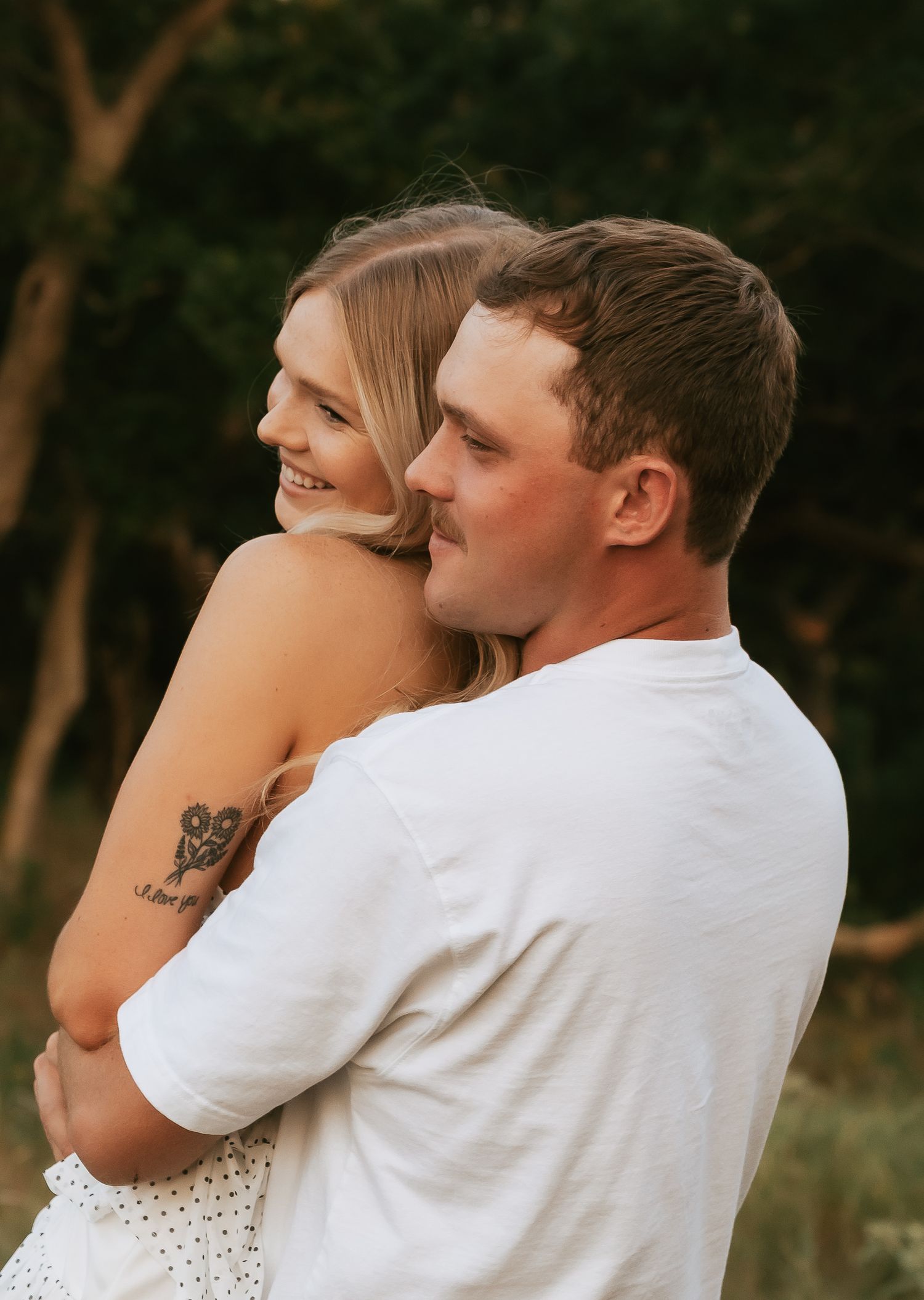 Couple embracing outdoors at golden hour, woman smiling with owl tattoo on arm, man in white t-shirt, trees in background.