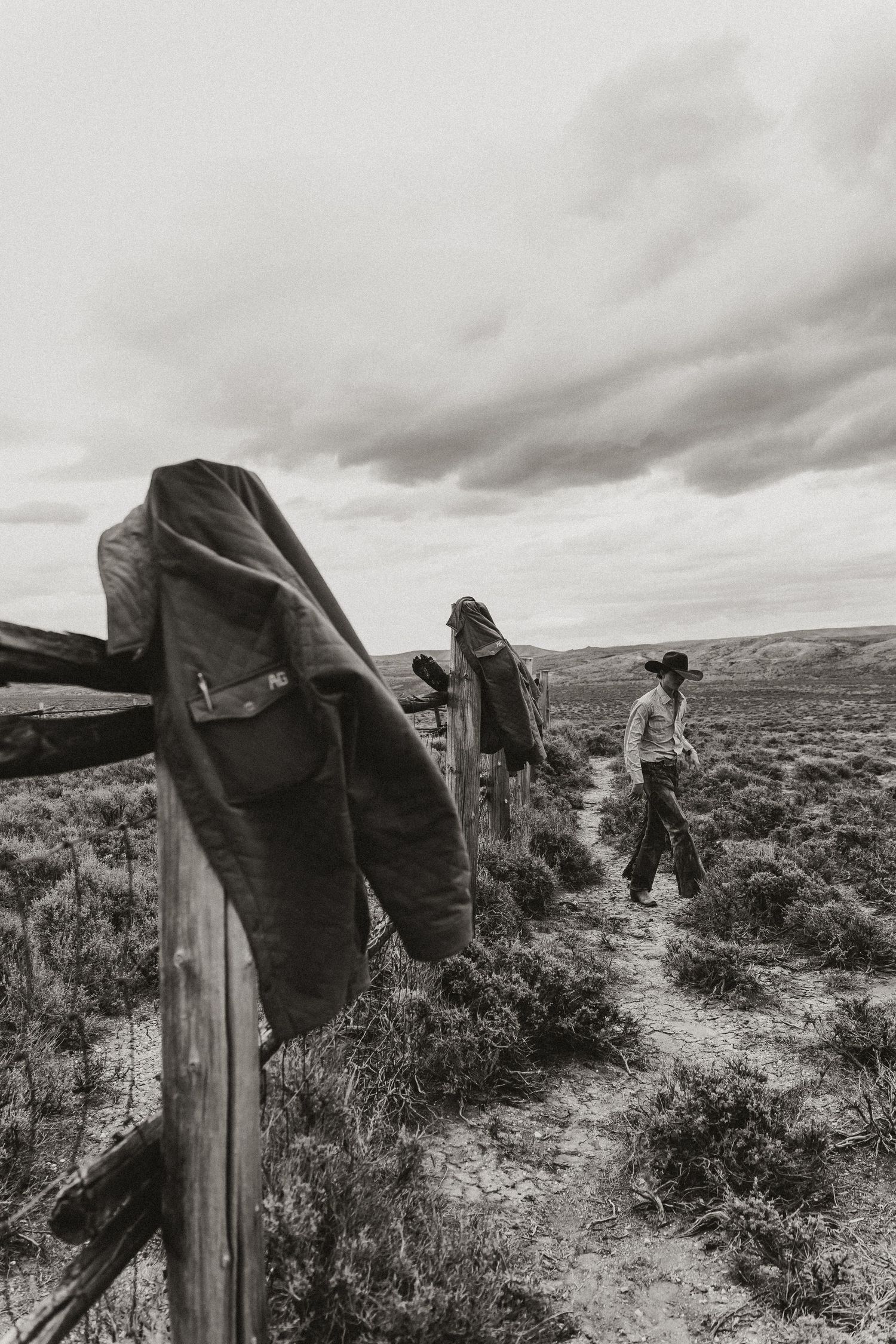 Black and white photo of a cowboy walking along a rustic fence in an open prairie landscape under cloudy skies.