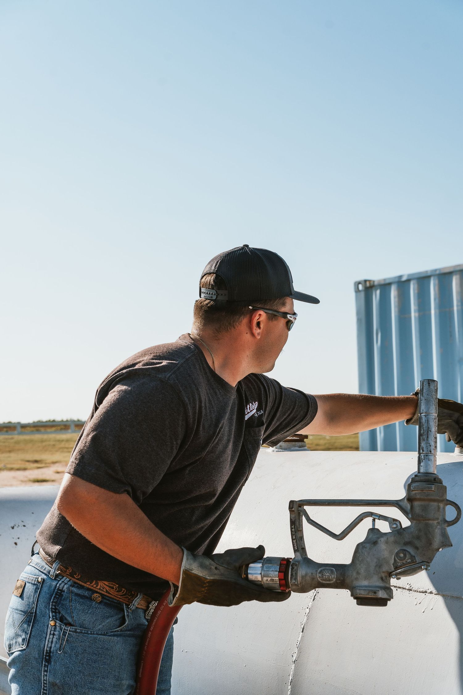 Worker in black cap and sunglasses operating fuel nozzle on white tanker truck outdoors.