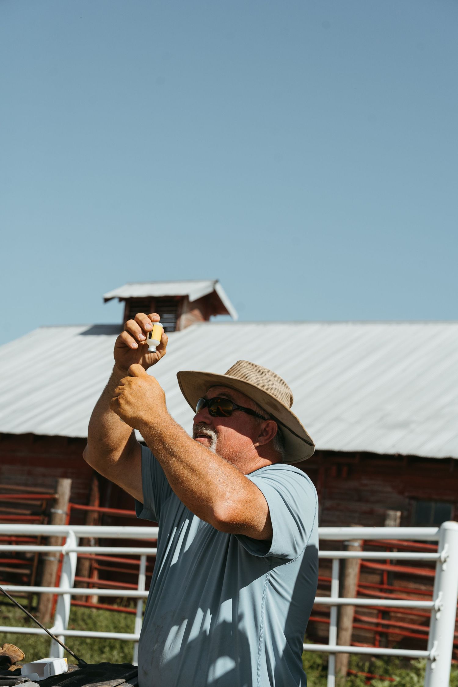 A man in a cowboy hat and sunglasses examines an object outdoors near a red barn fence on a sunny day.