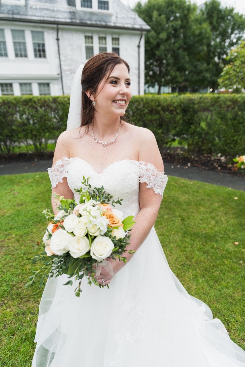 Beautiful bride in white off-shoulder gown holding elegant cream and peach floral bouquet.