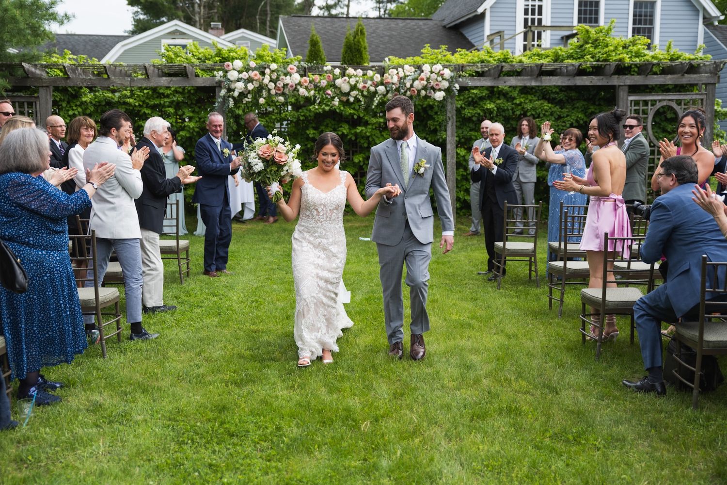 Wedding guests mingling in garden reception area with white house and trellis in background.