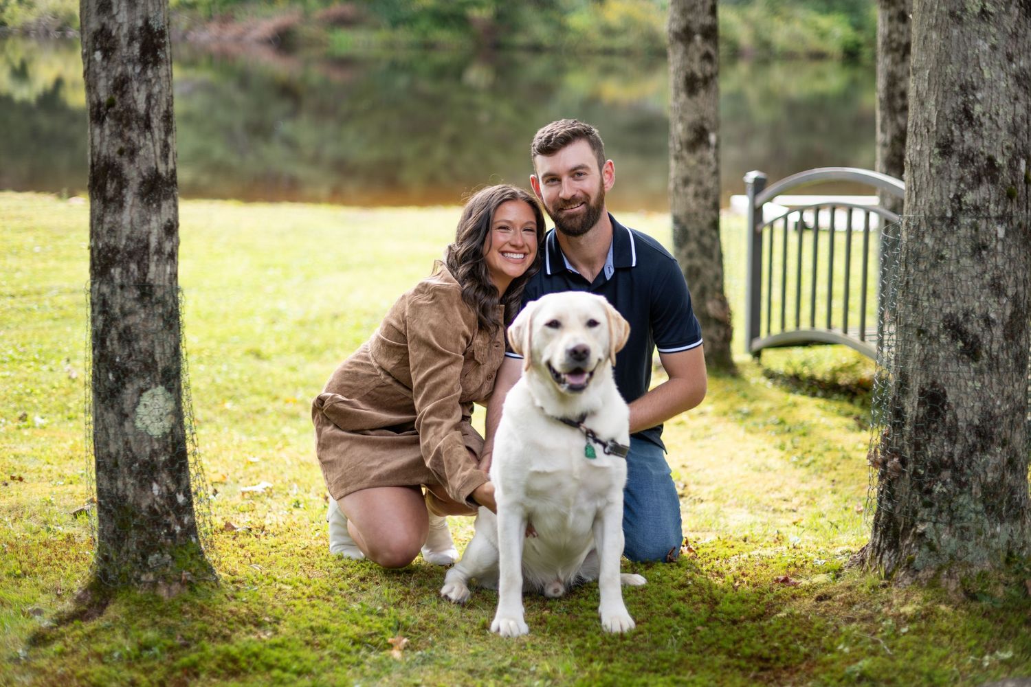 A Labrador retriever poses with couple under tree canopy during outdoor photoshoot.