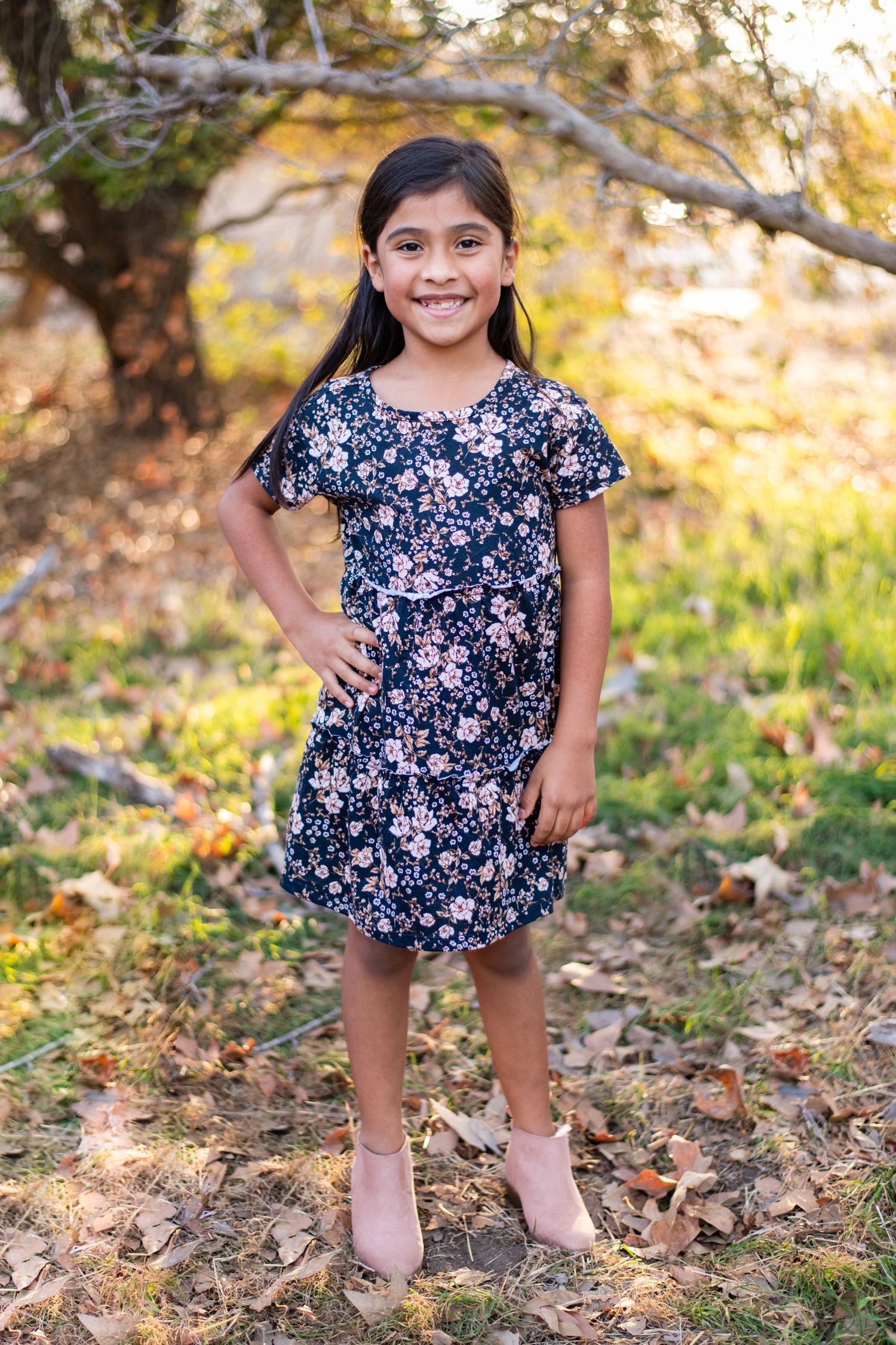 Little girl in a navy floral dress poses playfully among autumn leaves.