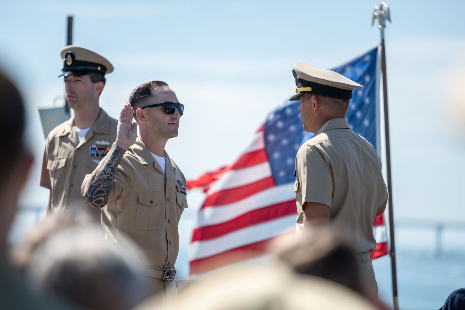 Military personnel stand at attention during a ceremony with an American flag flying in the background.