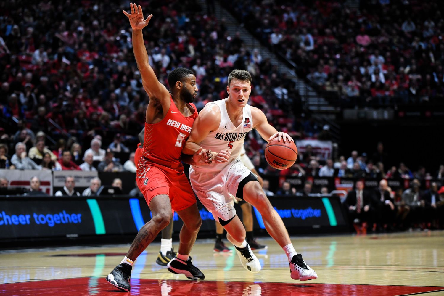 Two basketball players compete on court, one in white driving past a defender in red during an indoor game.