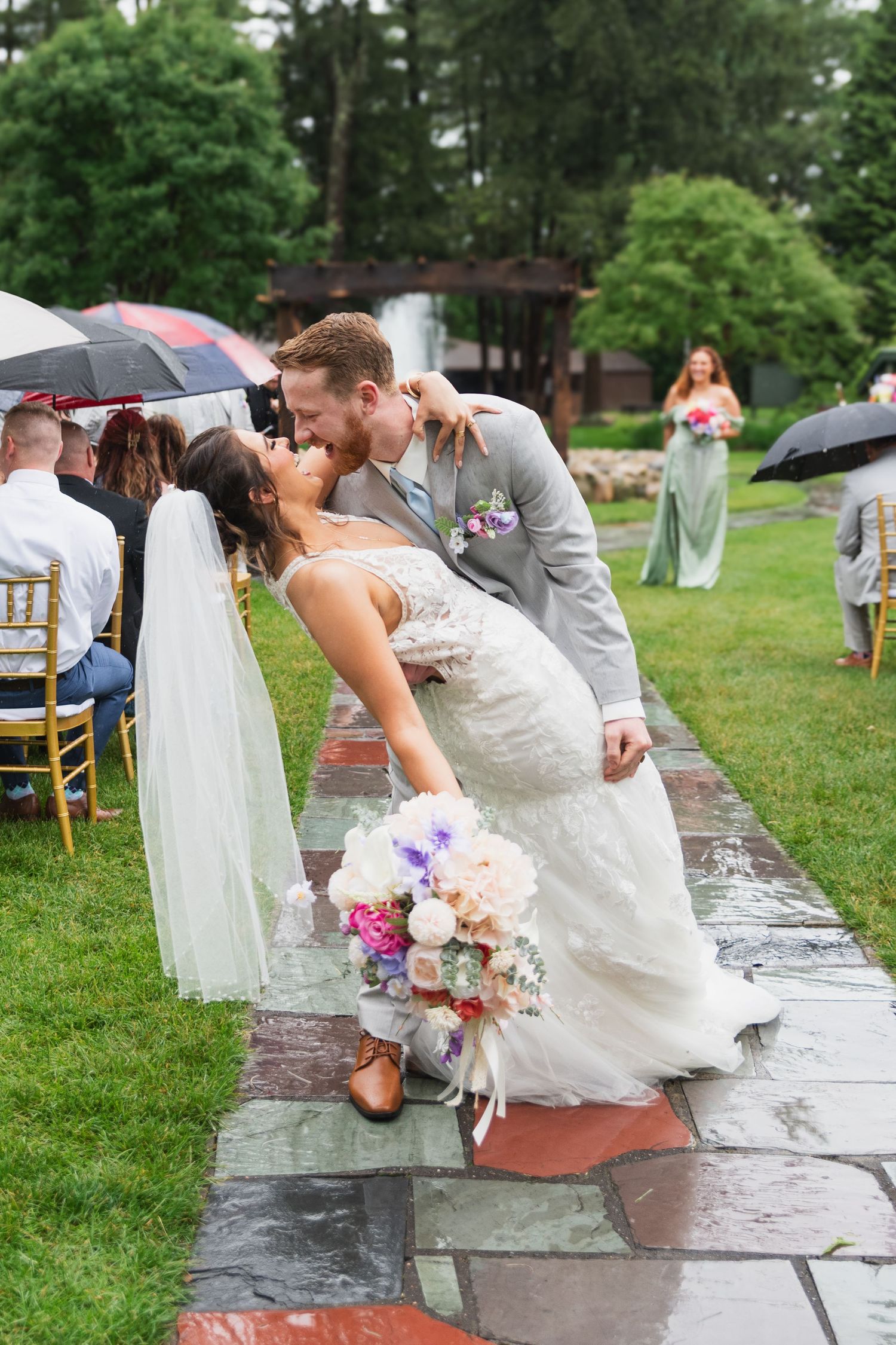 Wedding party members hop across wet stone path in garden during rainy outdoor ceremony.
