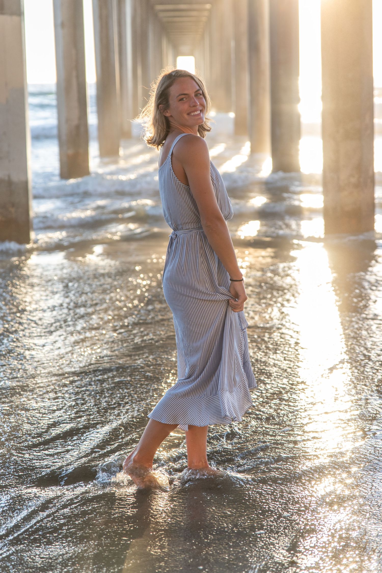 Woman in flowing gray dress standing under a pier at sunset as waves splash around her feet.