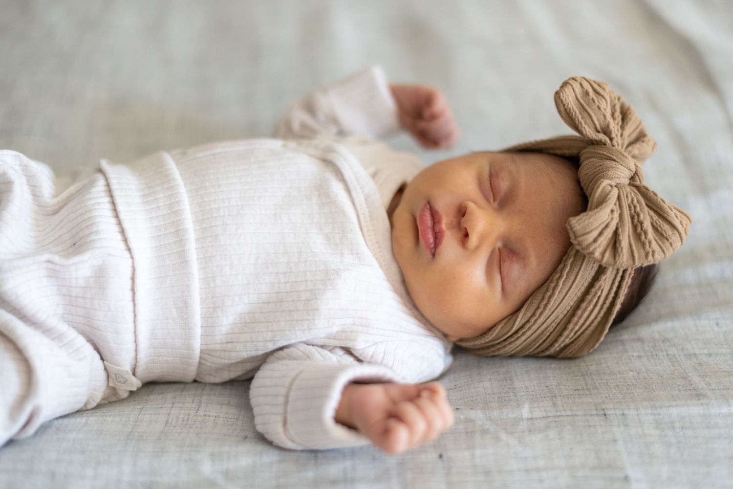 Sleeping baby wears neutral outfit with bow headband on light bedding.