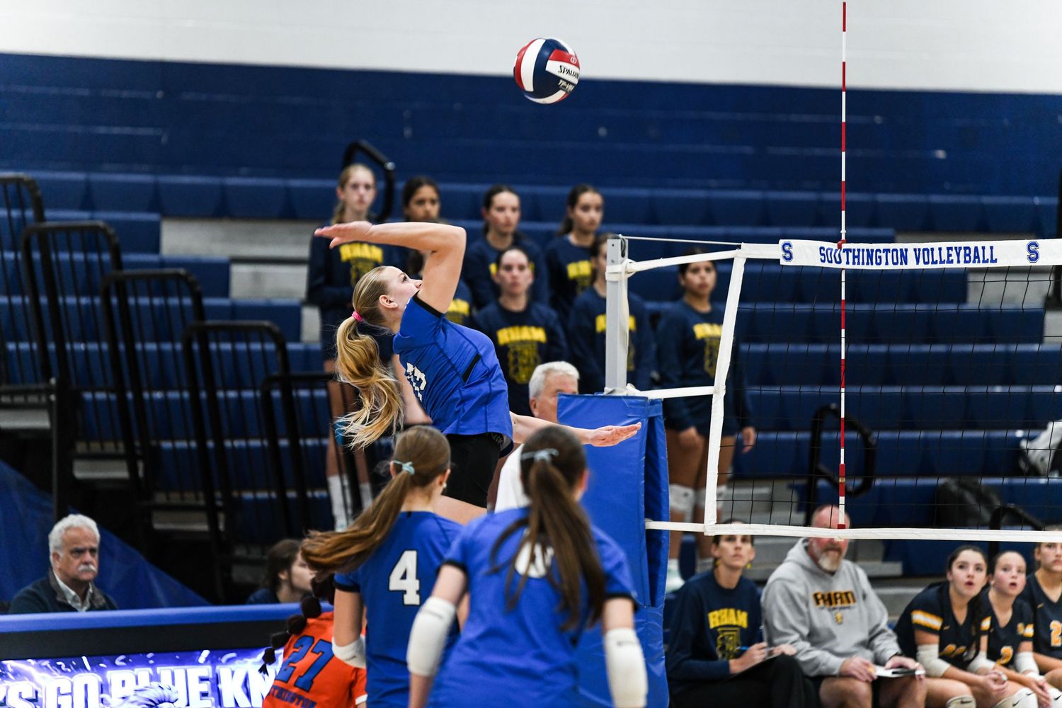 A female volleyball player in blue jumps to spike the ball over the net during a high school volleyball match.