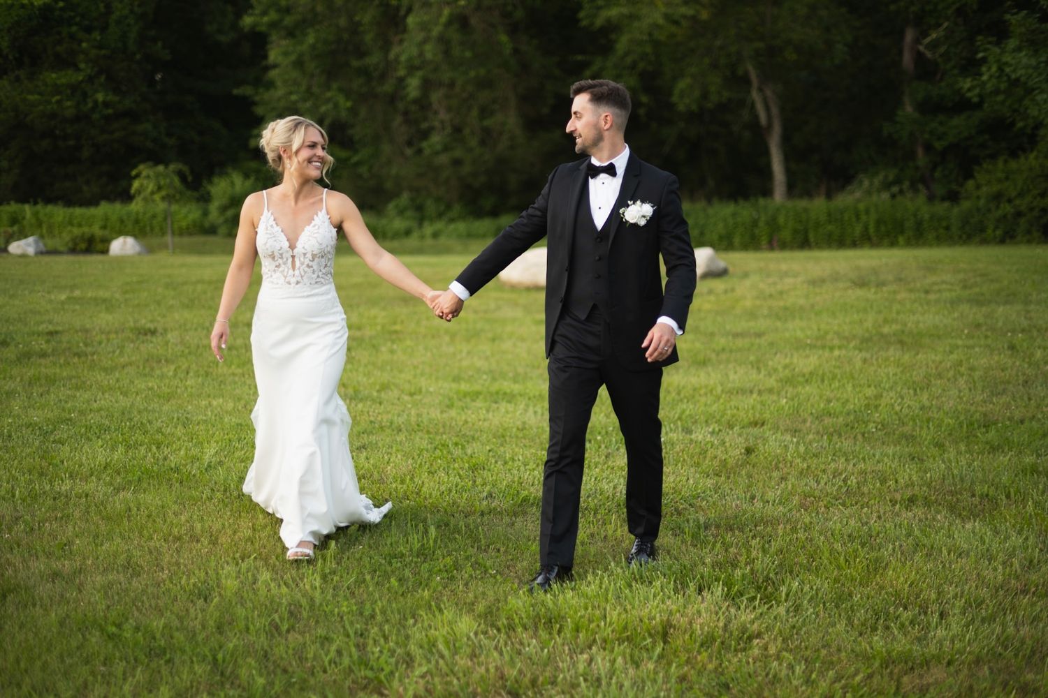 Newlyweds playfully run and dance together across a sunny green field.