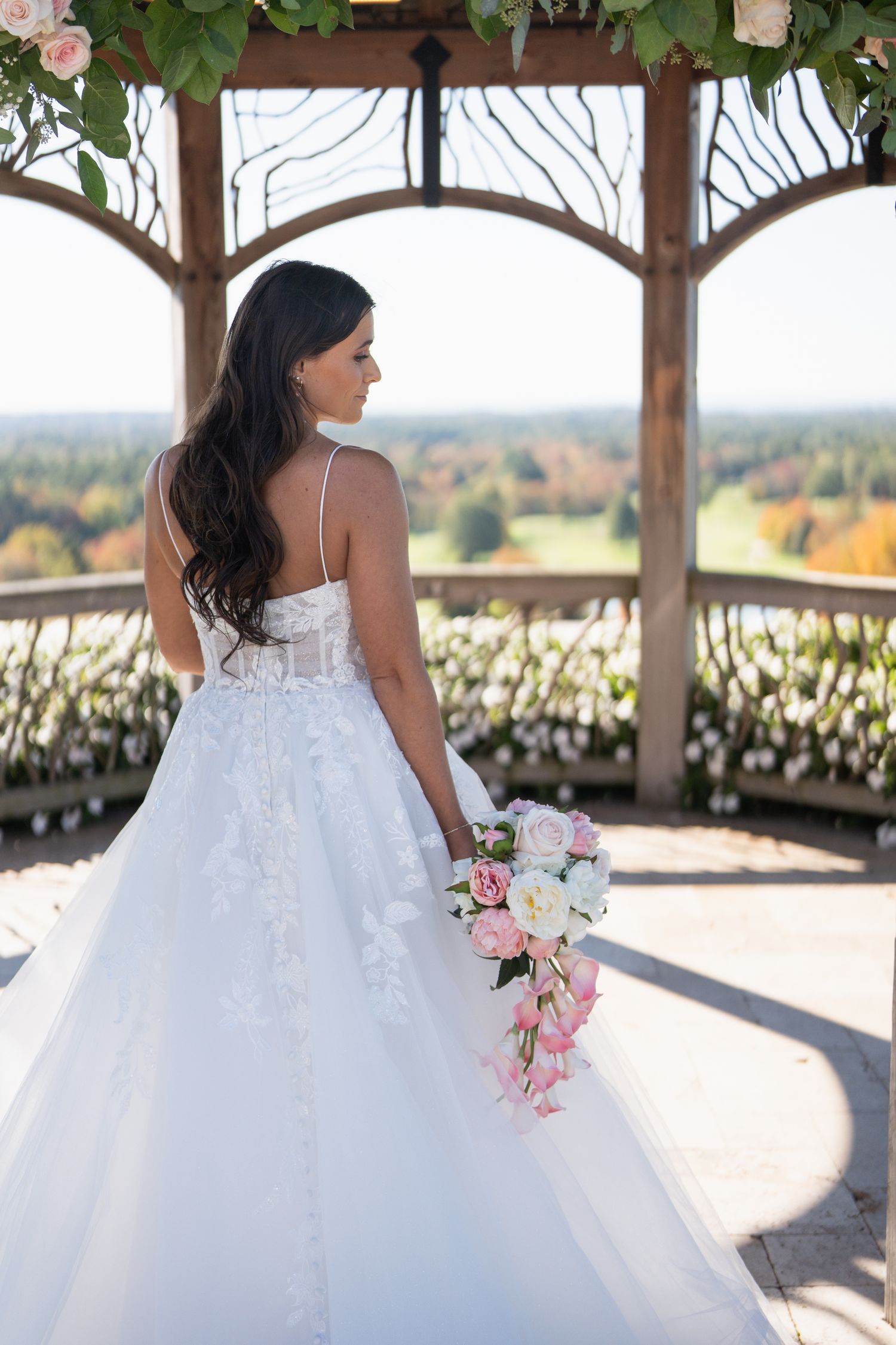 A bride in a white ballgown wedding dress sits on a rustic wooden bench holding a pink and white bouquet.