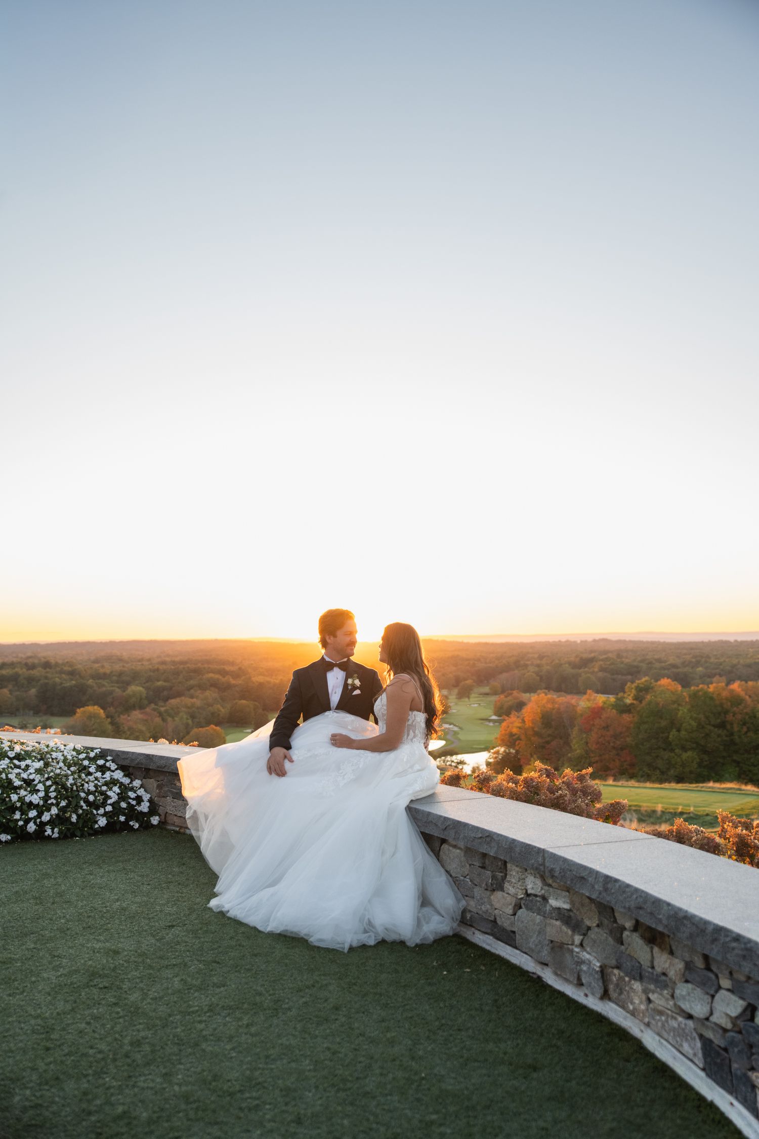 Couple in wedding attire sits on stone wall overlooking scenic countryside view at golden sunset.