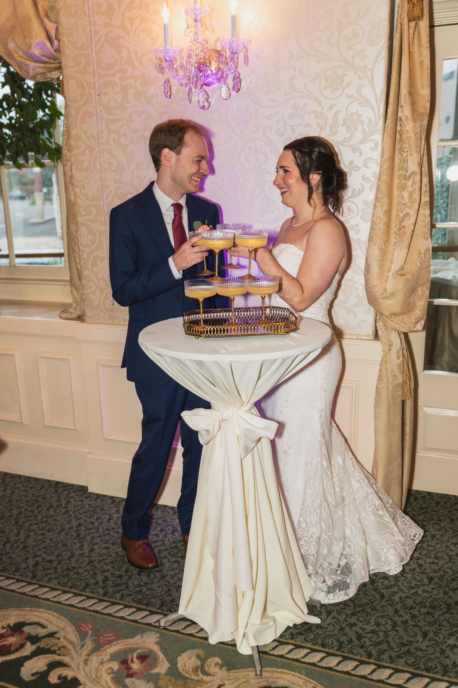 Newlyweds share cocktails at an elegant reception under a pink crystal chandelier.