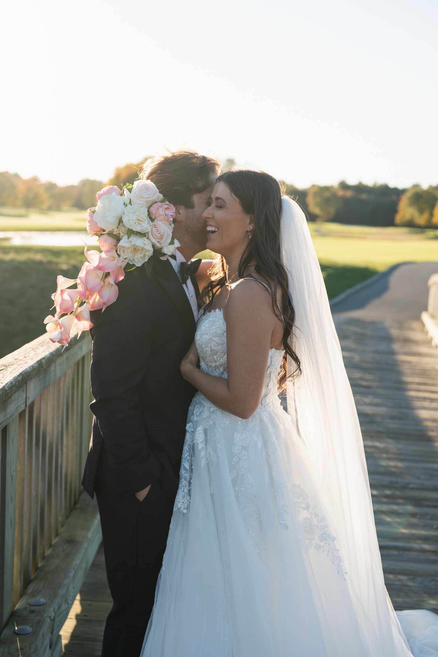 Newlyweds embrace on a wooden bridge during golden hour with pink roses in the background.