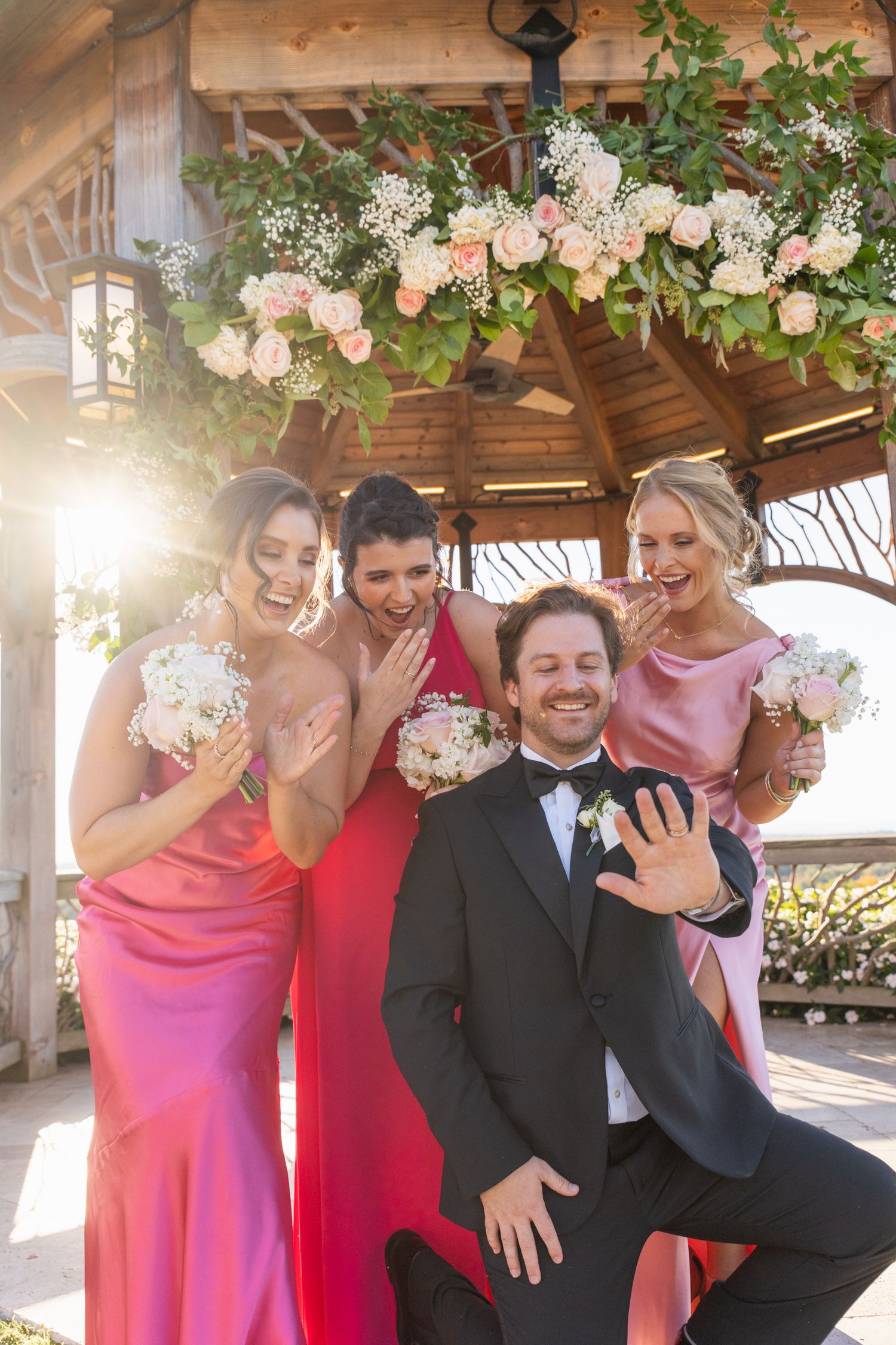 Wedding party wearing pink dresses and dark suits pose together under a wooden gazebo with floral decorations.
