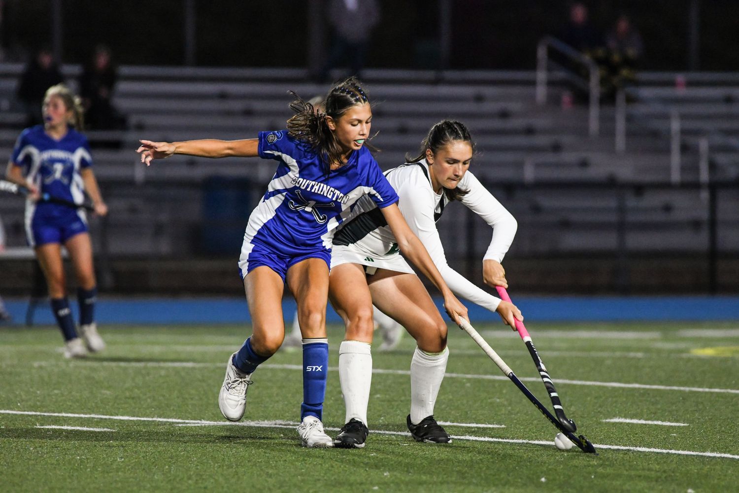 Two field hockey players compete for the ball during a night game, one in blue and one in white uniform.