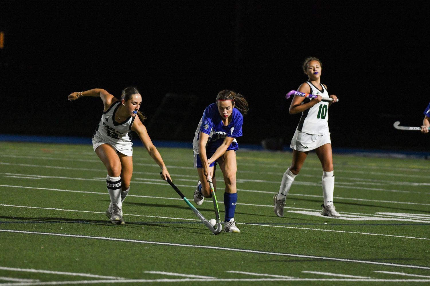 Female field hockey players compete in a night game, with one player in blue controlling the ball on a turf field.