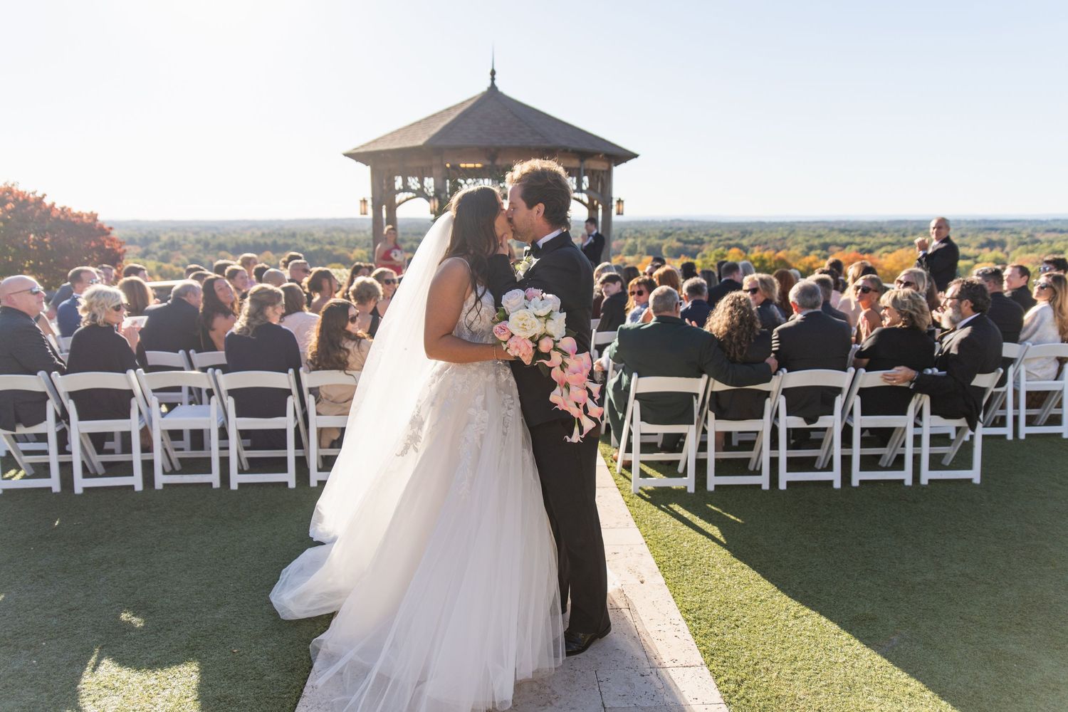 A wedding ceremony setup on a hilltop overlooks a scenic valley with white chairs arranged in rows at sunset.