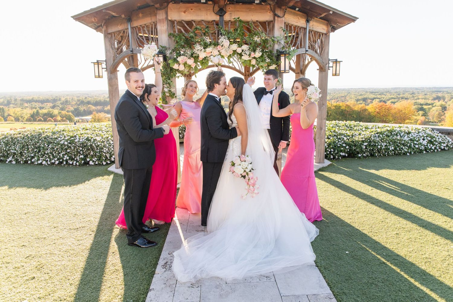 Wedding party gathered at an outdoor gazebo with floral decor overlooking scenic countryside on sunny afternoon.