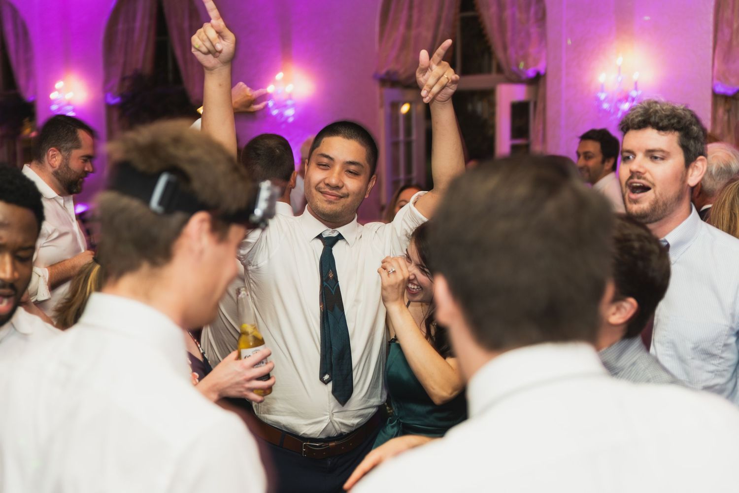 Wedding guests dancing and celebrating on a dance floor illuminated with purple lighting at a reception party.