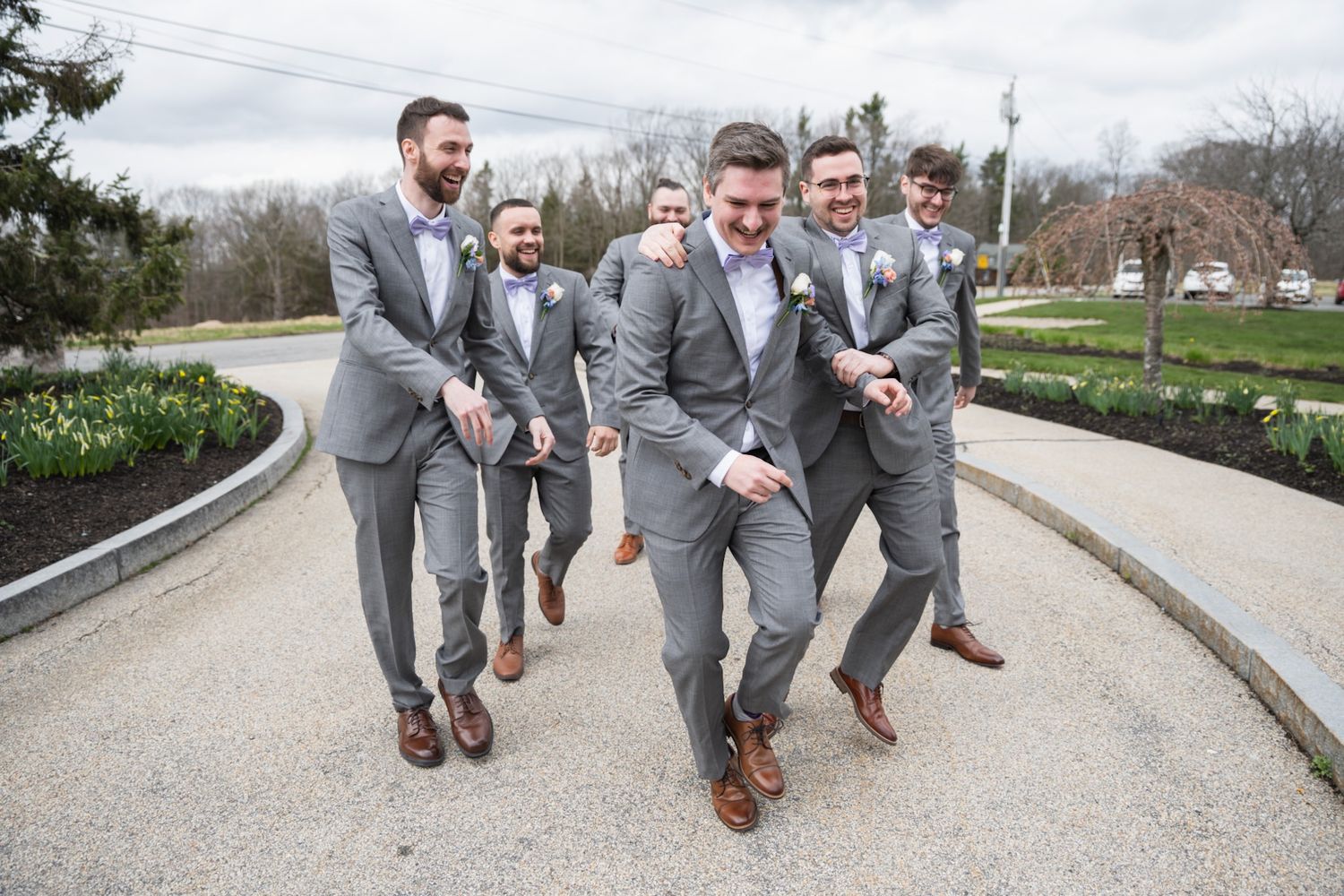 Groomsmen in gray suits jump and pose on concrete walkway.
