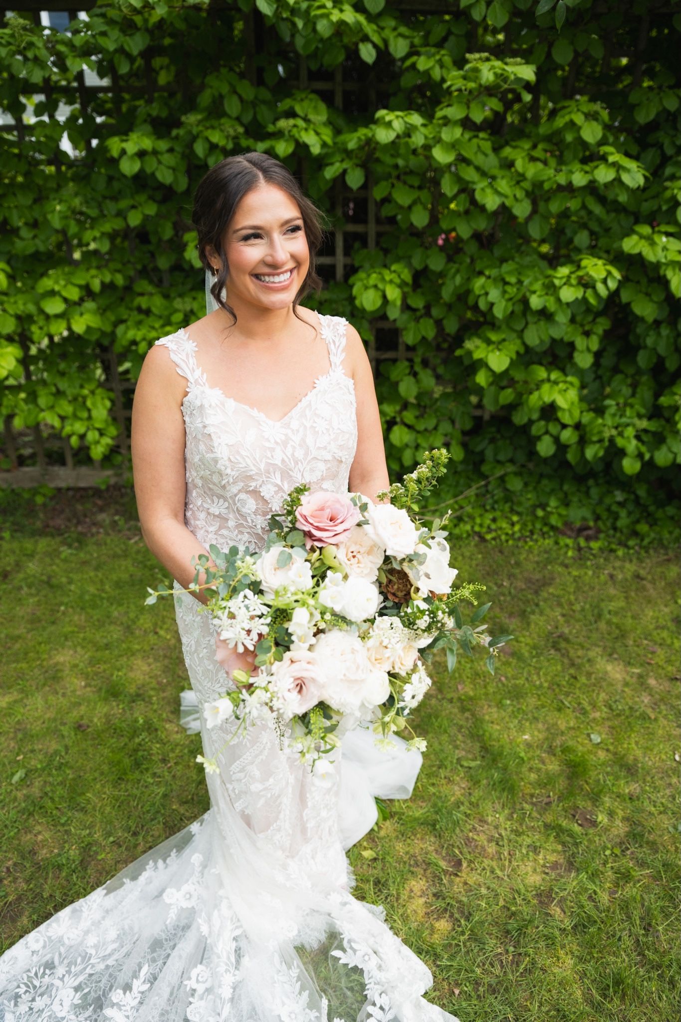 Bride in lace dress holds a beautiful white and pink floral bouquet against green foliage.