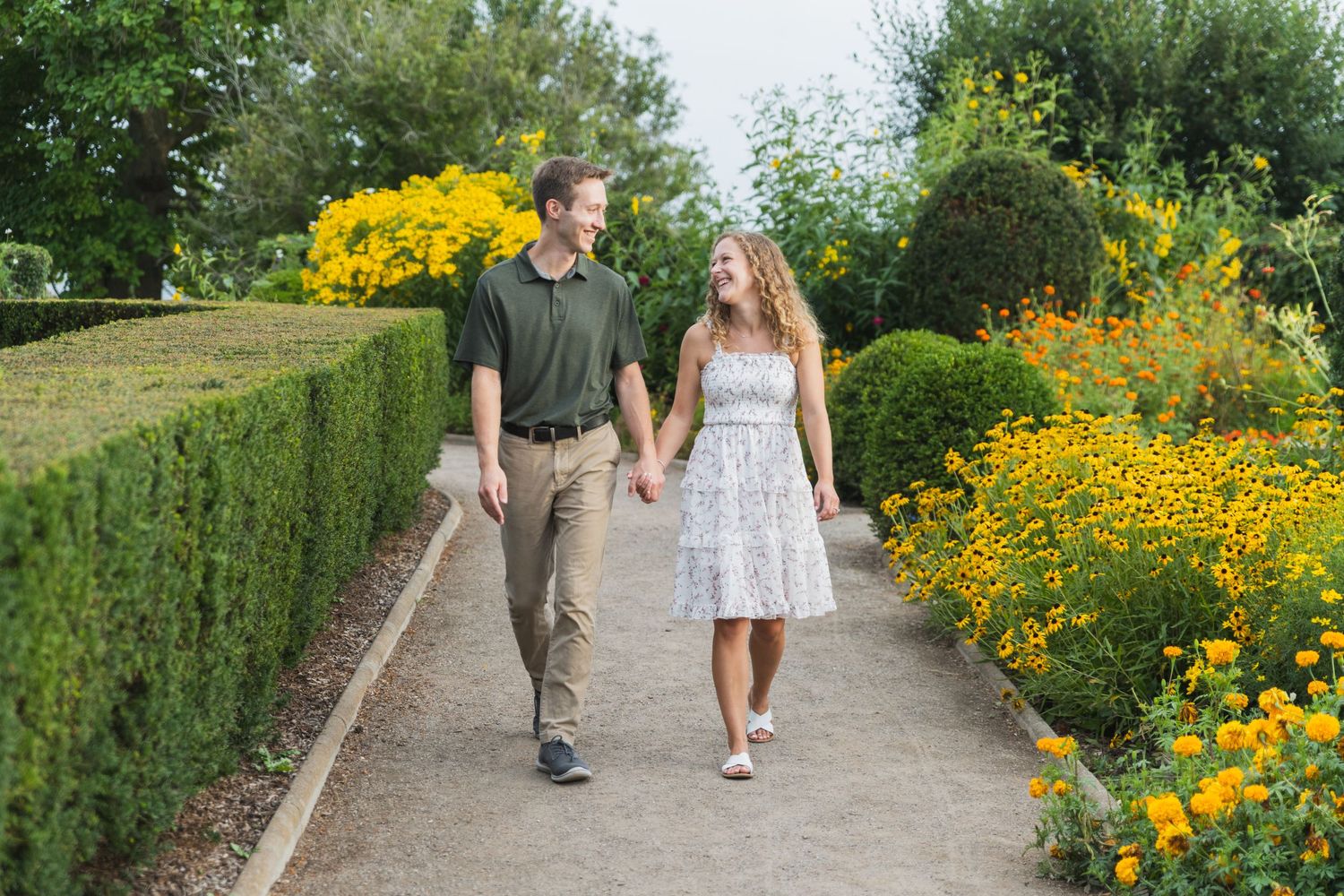 A couple walks hand in hand along a garden pathway with vibrant yellow flowers and lush greenery surrounding them.